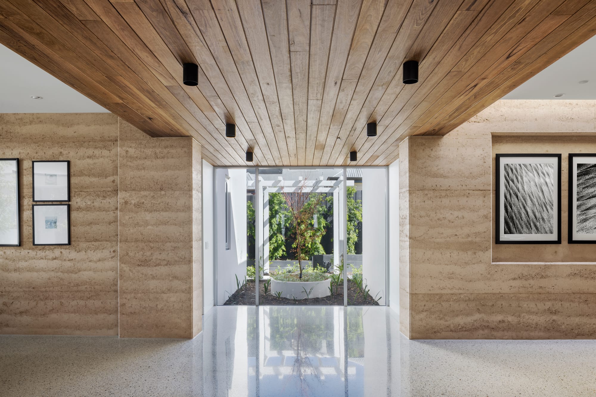 Earth + Sky House by Kirsten Johnstone Architecture. Photography by Tatjana Plitt. Interior with polished concrete flooring, rammed earth walls and timber clad ceiling, with glass windows overlooking into courtyard with round planter with tree in it. 