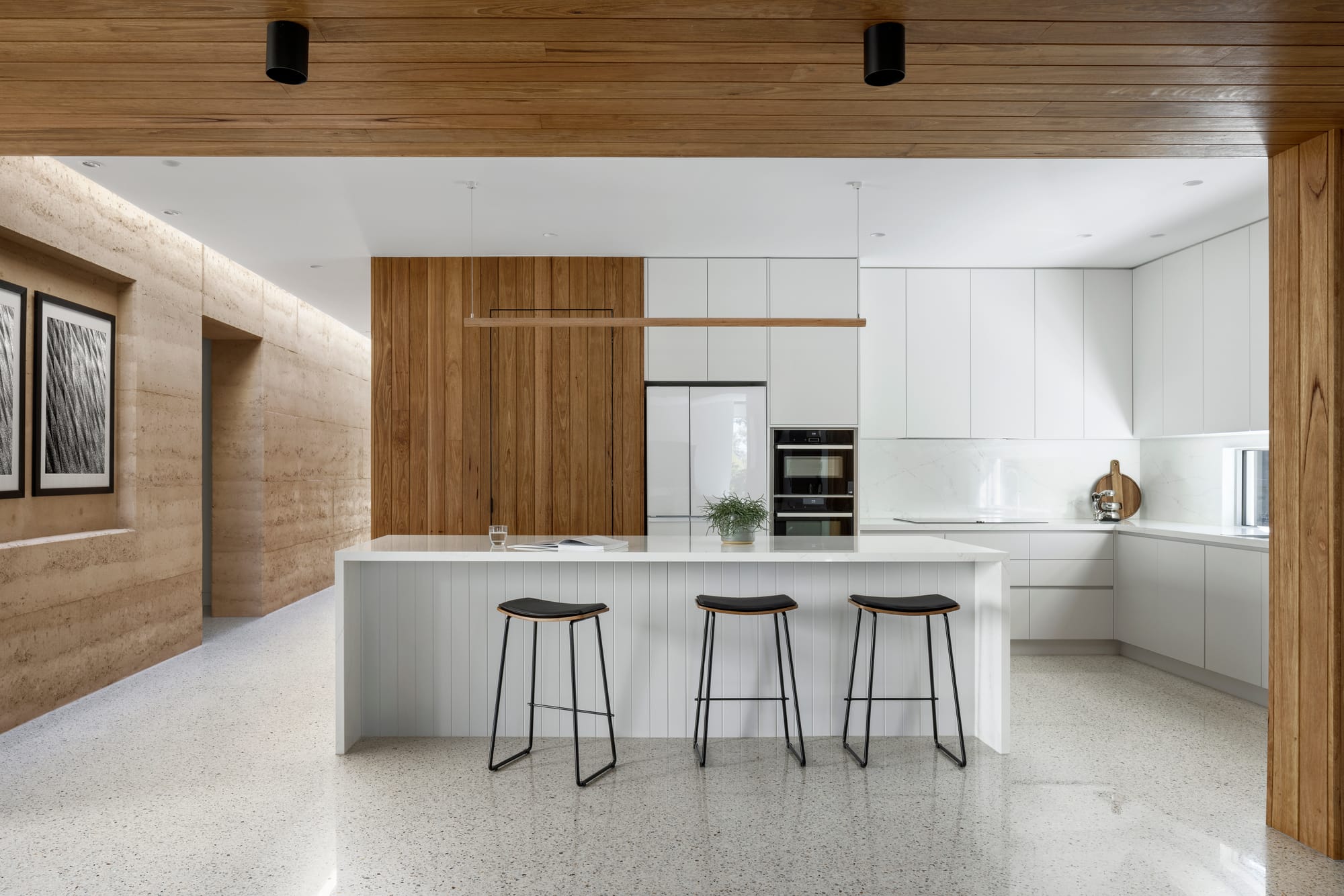 Earth + Sky House by Kirsten Johnstone Architecture. Photography by Tatjana Plitt. Kitchen with polished concrete flooring, white cabinetry, timber walls and ceilings, and black bar stools. 