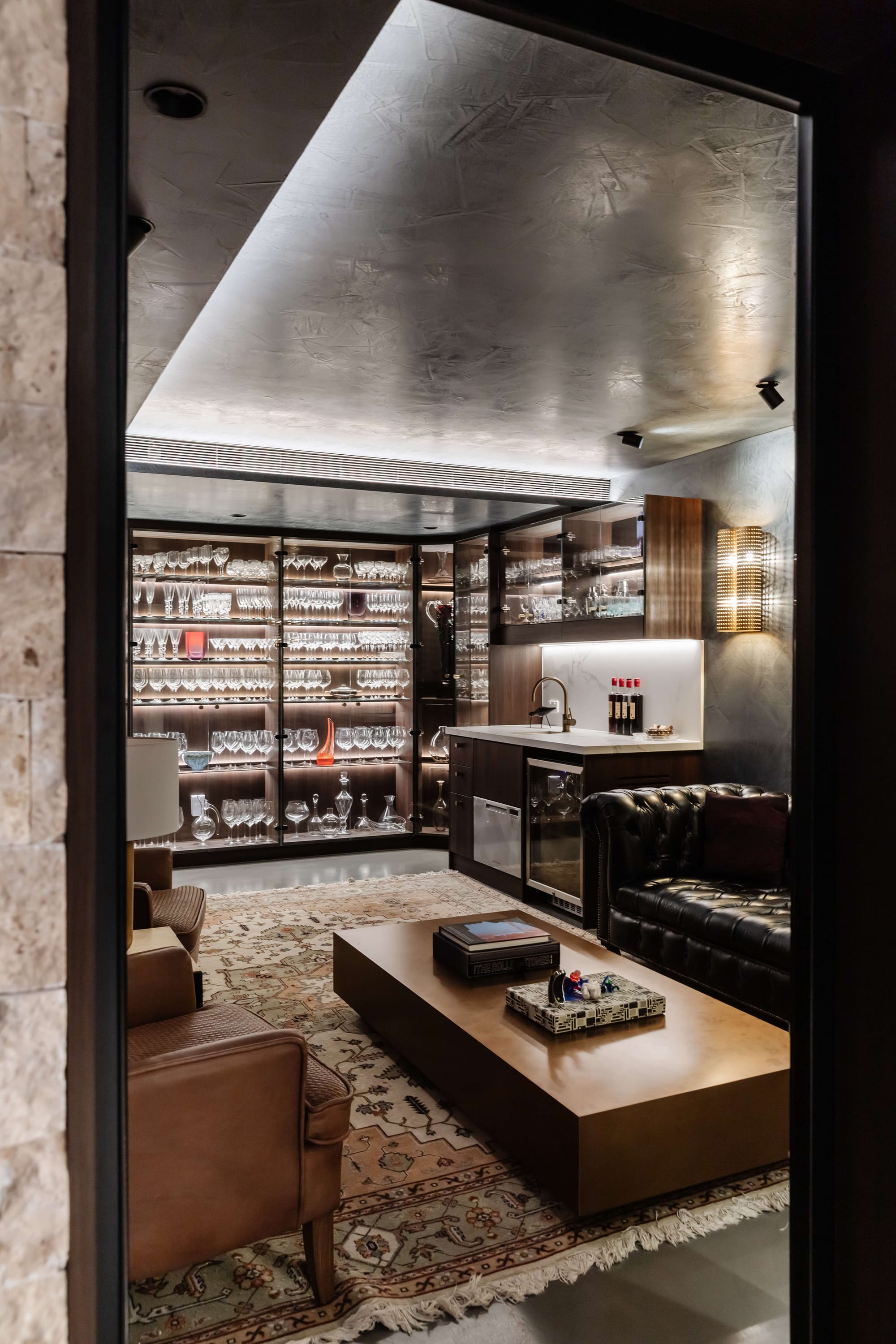 Avalon Beach Wine Cellar & Lounge by Nx Studio. Photography by REM Design House. Seating area with black leather couch, brown leather armchair, brass coffee table, patterned rug and large timber storage units with glassware.