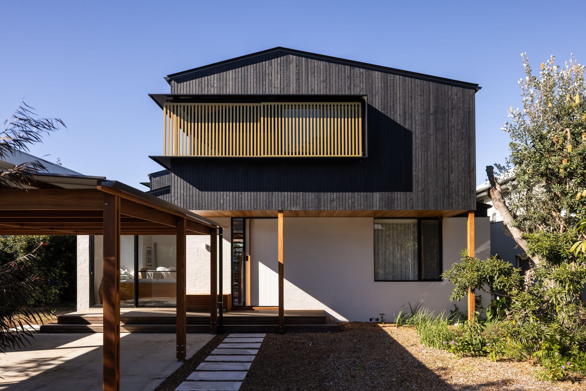 Double storey home with white brick lower-level facade, charcoal timber clad upper level, and grey stone pavers through mulch to timber deck.   