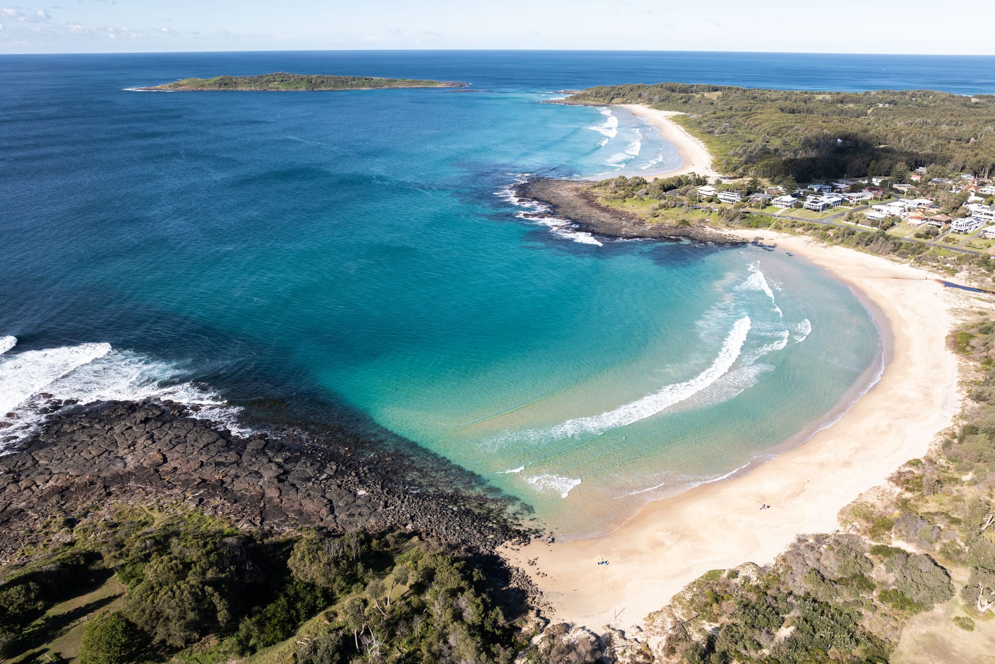 Bawley Point by Watershed Architects. Photography by Simon Whitbread. Aerial view of coastline with dense foliage, rocky shoreline and small township to left of beach. 