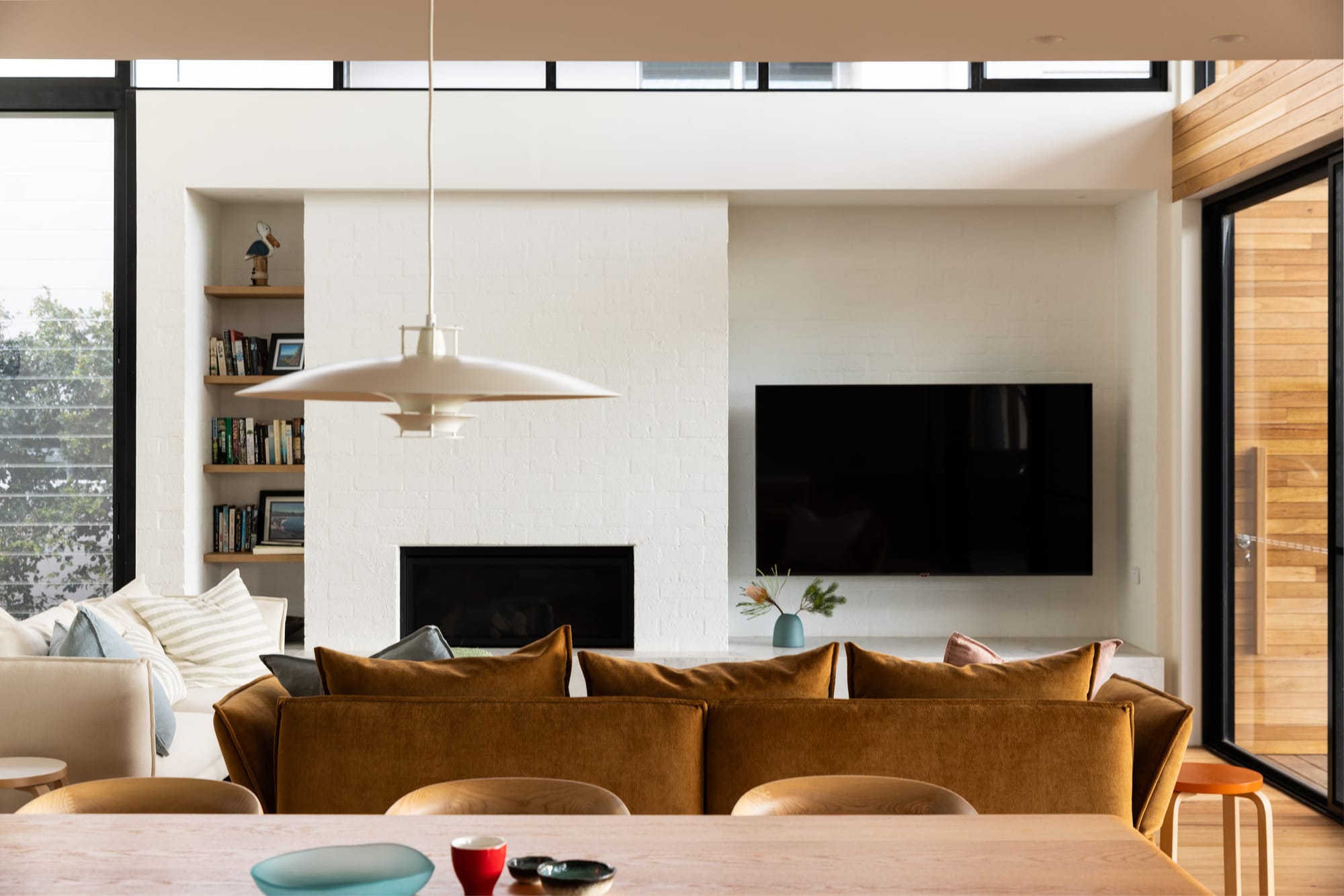 Bawley Point by Watershed Architects. Photography by Simon Whitbread. Living space with suede couch, white brick wall with integrated fireplace and television, white pendant lighting and large overhead void. 
