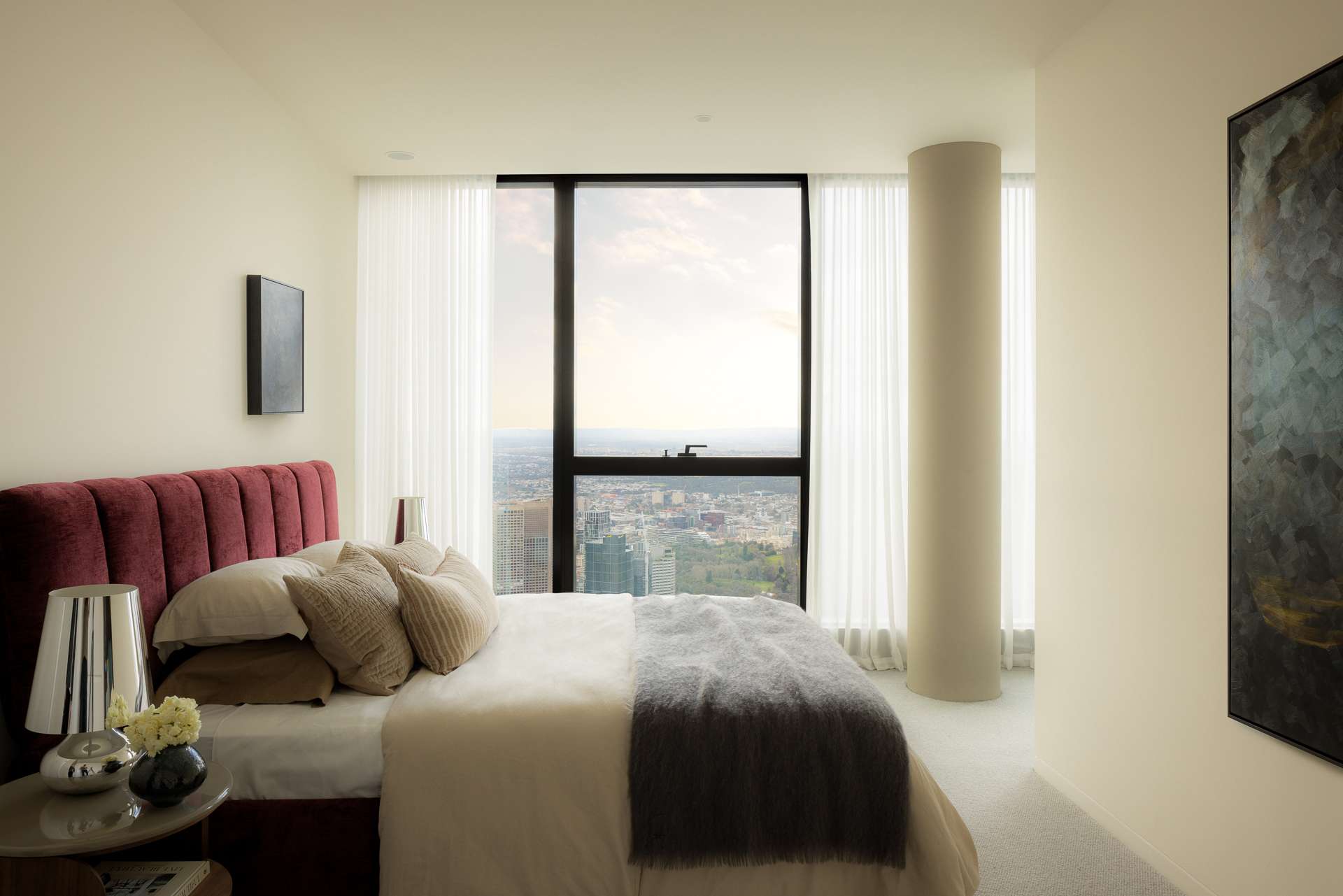 Cloud Residence by FURNISHD. Photography by Dylan James. Bedroom with white walls, dark red headboard, full height windows with city views, white sheers and grey throw rug on bed. 