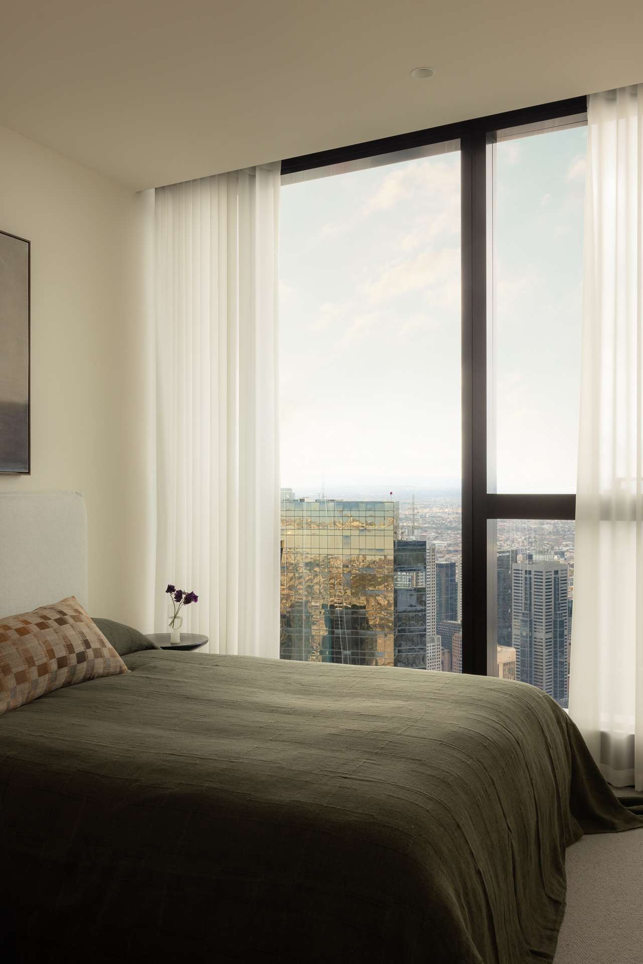 Cloud Residence by FURNISHD. Photography by Dylan James. Bedroom with olive bedding, full height glass windows with city skyline views, and white sheers. 