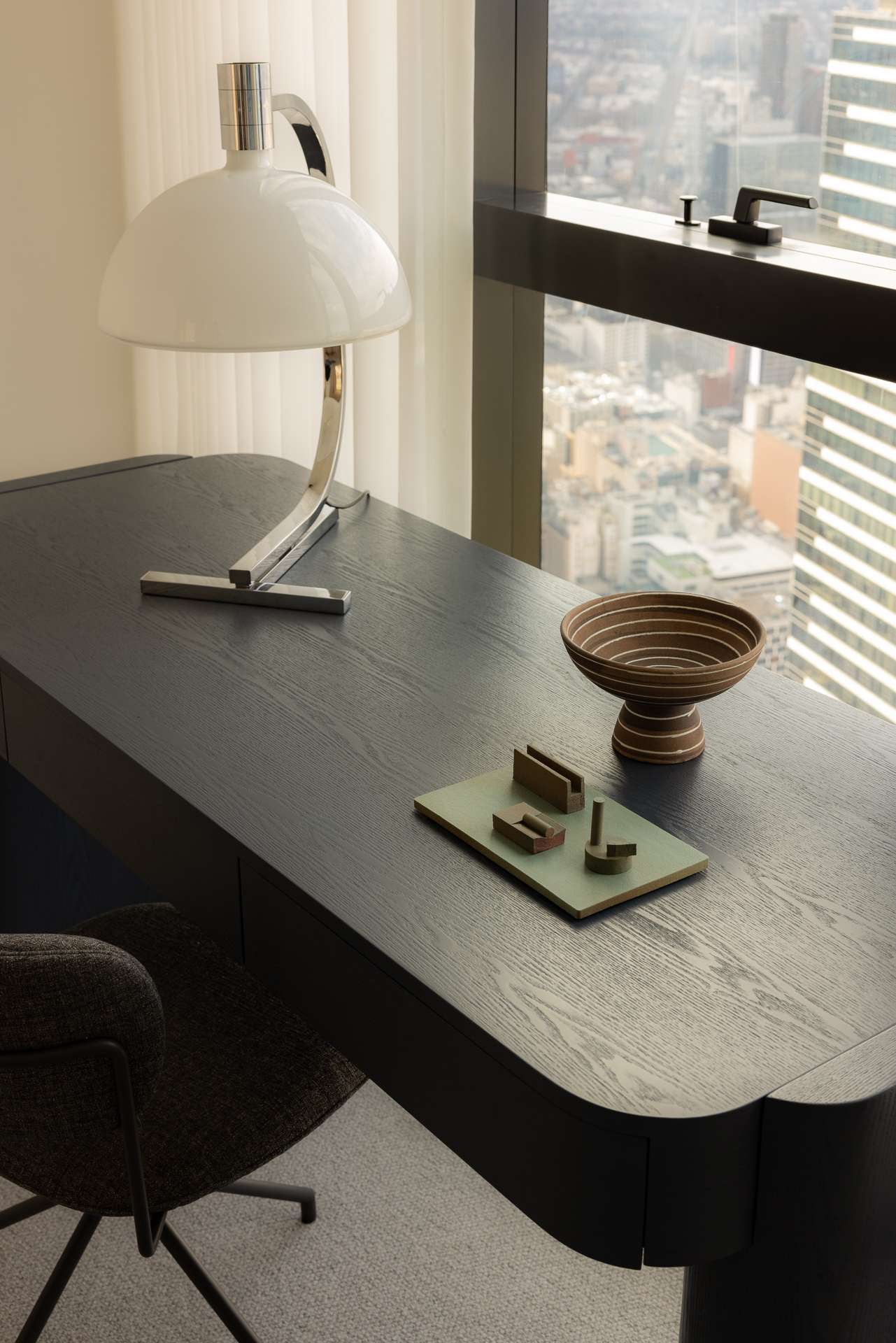 Cloud Residence by FURNISHD. Photography by Dylan James. Close up of black timber deck with chrome lamp and lack office chair, pushed against windows overlooking city from above. 