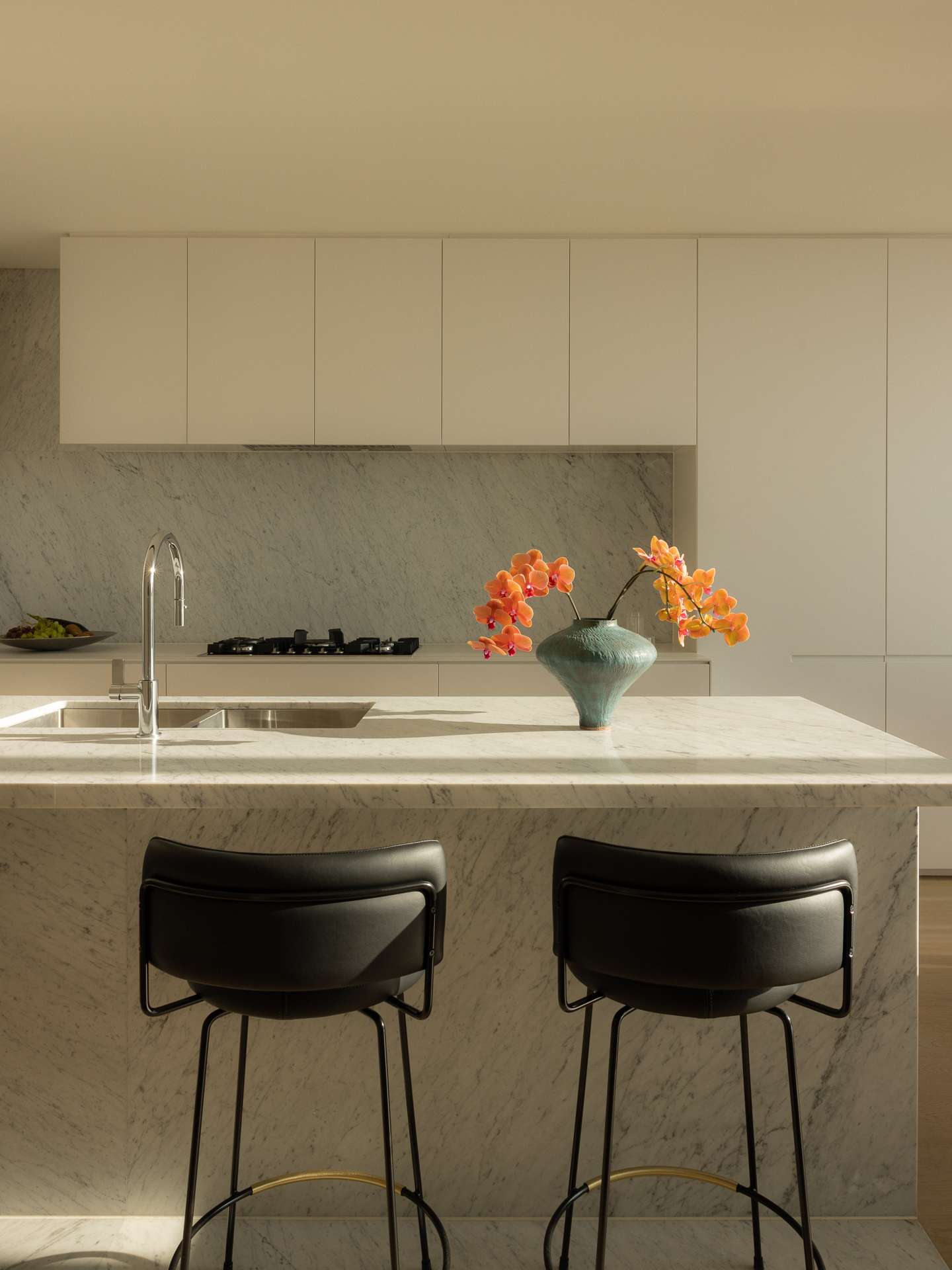 Cloud Residence by FURNISHD. Photography by Dylan James. Kitchen with white cabinets, white stone island bench, countertops and splashbacks, and black bar stools. 