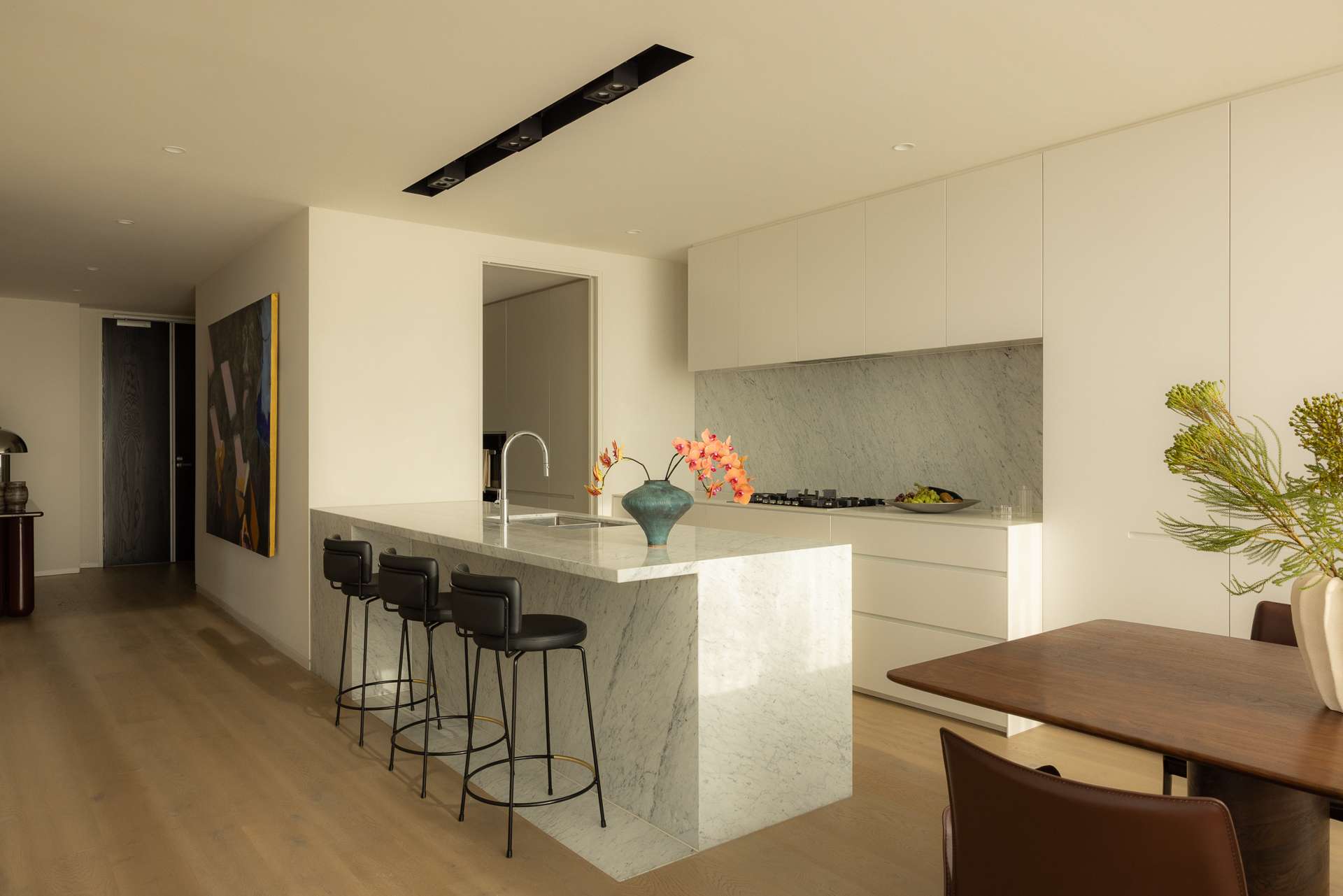 Cloud Residence by FURNISHD. Photography by Dylan James. Kitchen and dining space with light timber flooring, rich timber dining table, white stone island bench, countertops and splashback, and white cabinetry. 