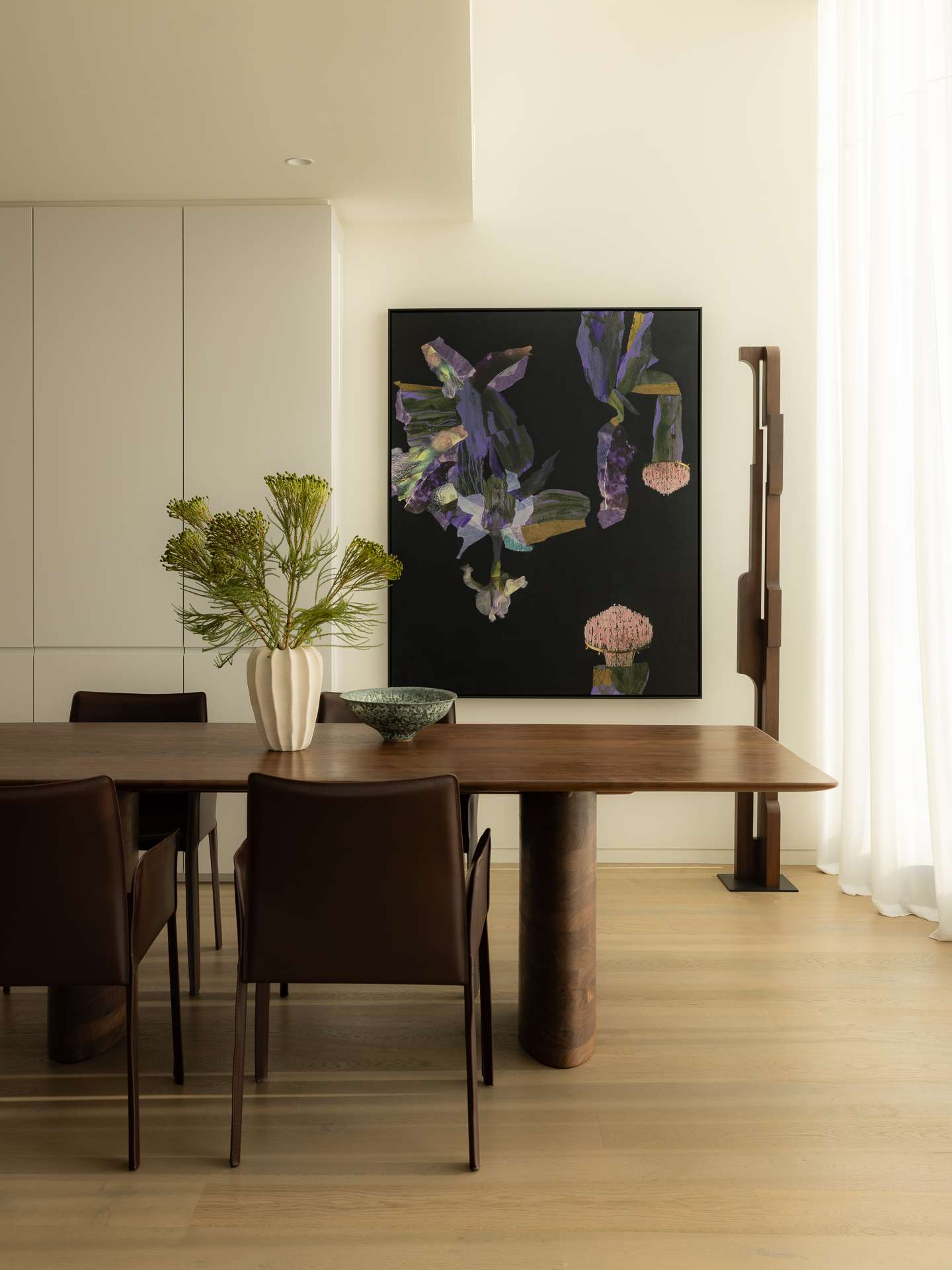Cloud Residence by FURNISHD. Photography by Dylan James. Dark timber dining table with chocolate brown leather chairs, positioned in front of abstract wall art in background, white cabinets to left and white sheers to right. 