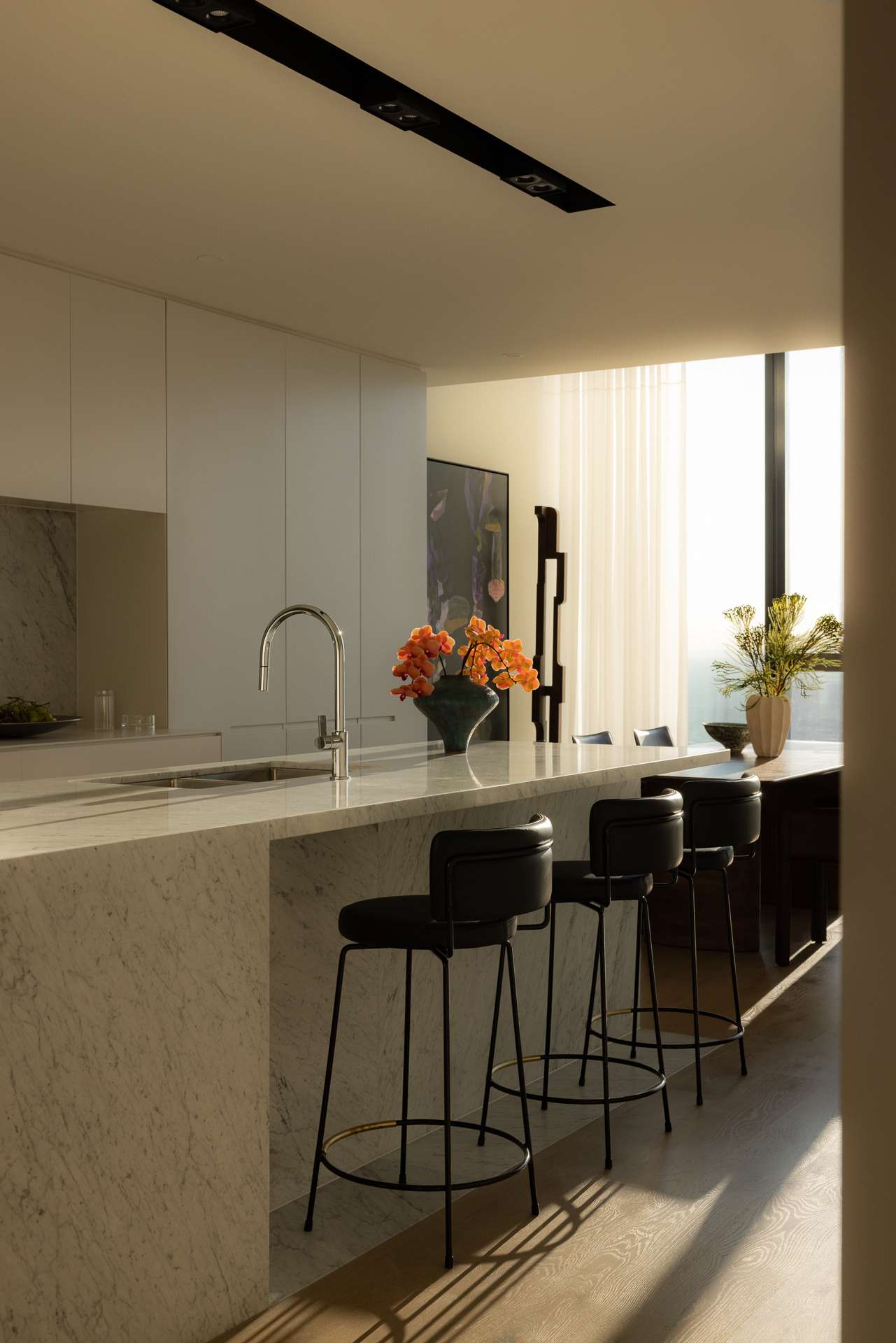 Cloud Residence by FURNISHD. Photography by Dylan James. Sunlight streaming through full height window to right of image, across kitchen with stone island, white cabinetry and black stools. 