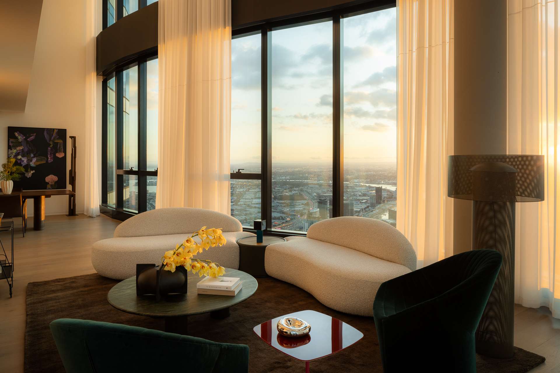 Cloud Residence by FURNISHD. Photography by Dylan James. Open plan living space with curved, floor-to-ceiling glass windows overlooking city, letting warm light into space across white couches. 