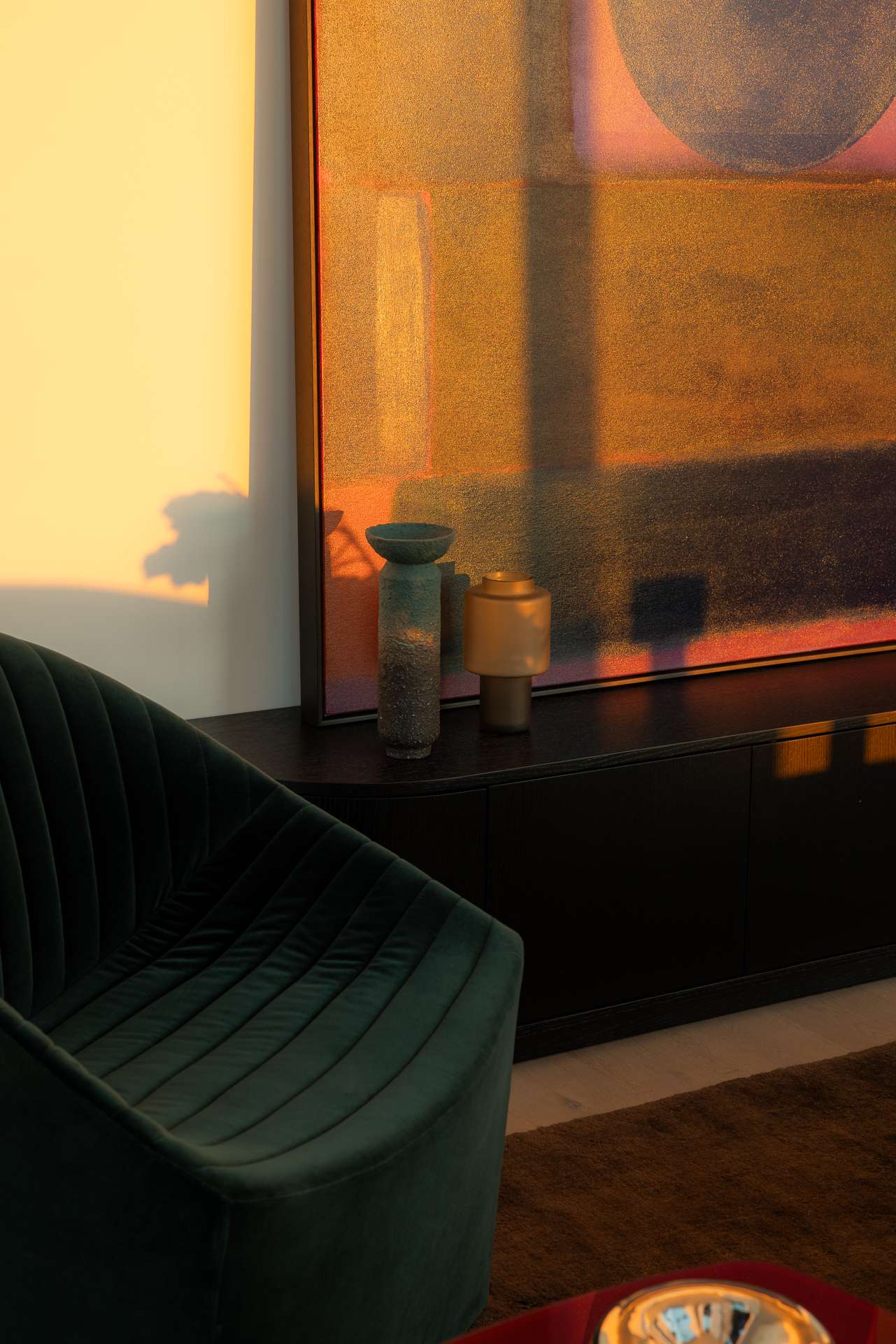 Cloud Residence by FURNISHD. Photography by Dylan James. Close up of dark green armchair and abstract art on black sideboard, with golden light casted across them. 