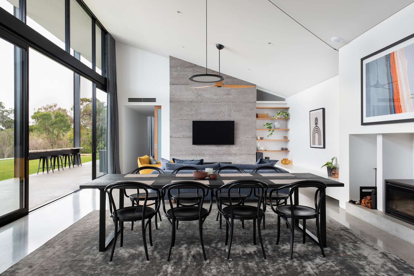 Hausen Court by Keen Architecture. Photography by Keen Architecture. Open plan dining and living space with angled white ceiling, full height glass windows, concrete feature wall, black dining table and grey velvet floor rug. 