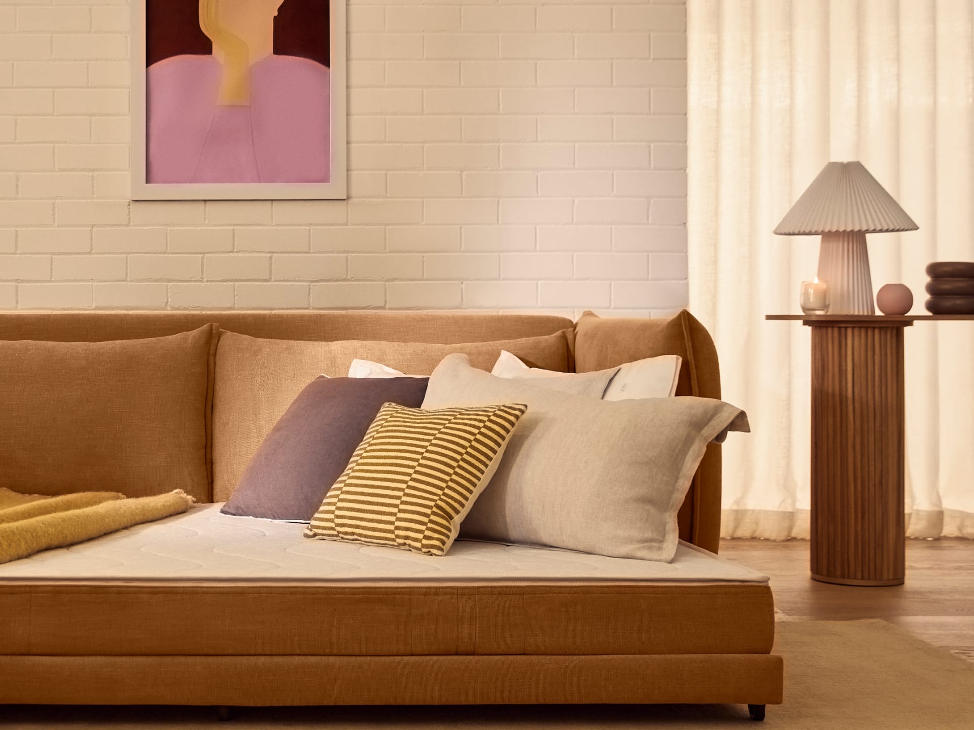 Koala Byron Sofa Bed [3rd Gen] in Bronzed Aussie. Fold out sofa bed in mustard colour, with purple, beige and yellow pills in front of white brick wall and sheers. 