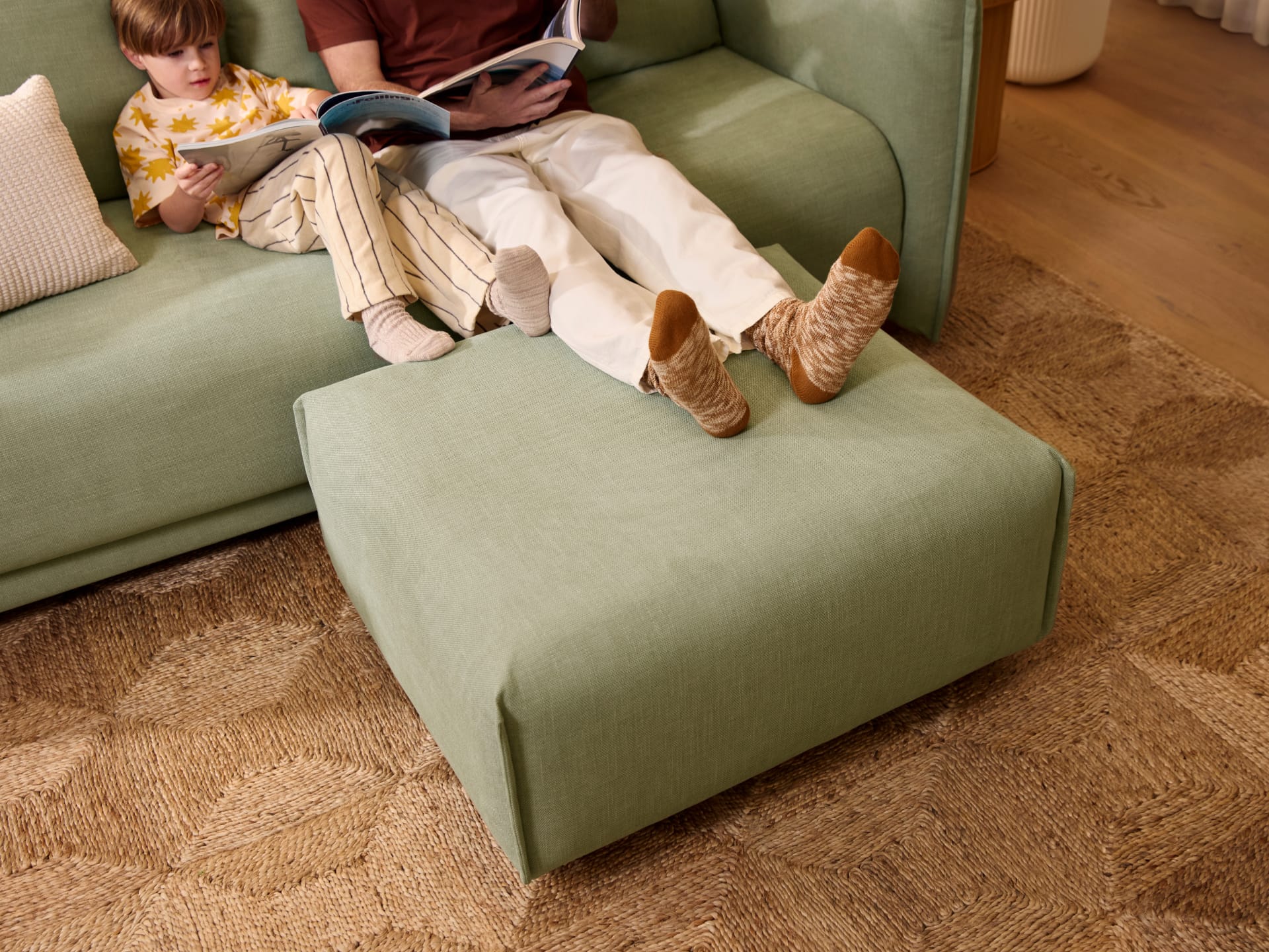 Koala Byron Sofa Bed [3rd Gen] in Cool Cactus. Light olive green couch with marching square ottoman on top of patterned jute rug, with one adult and one child resting their feet on ottoman. 