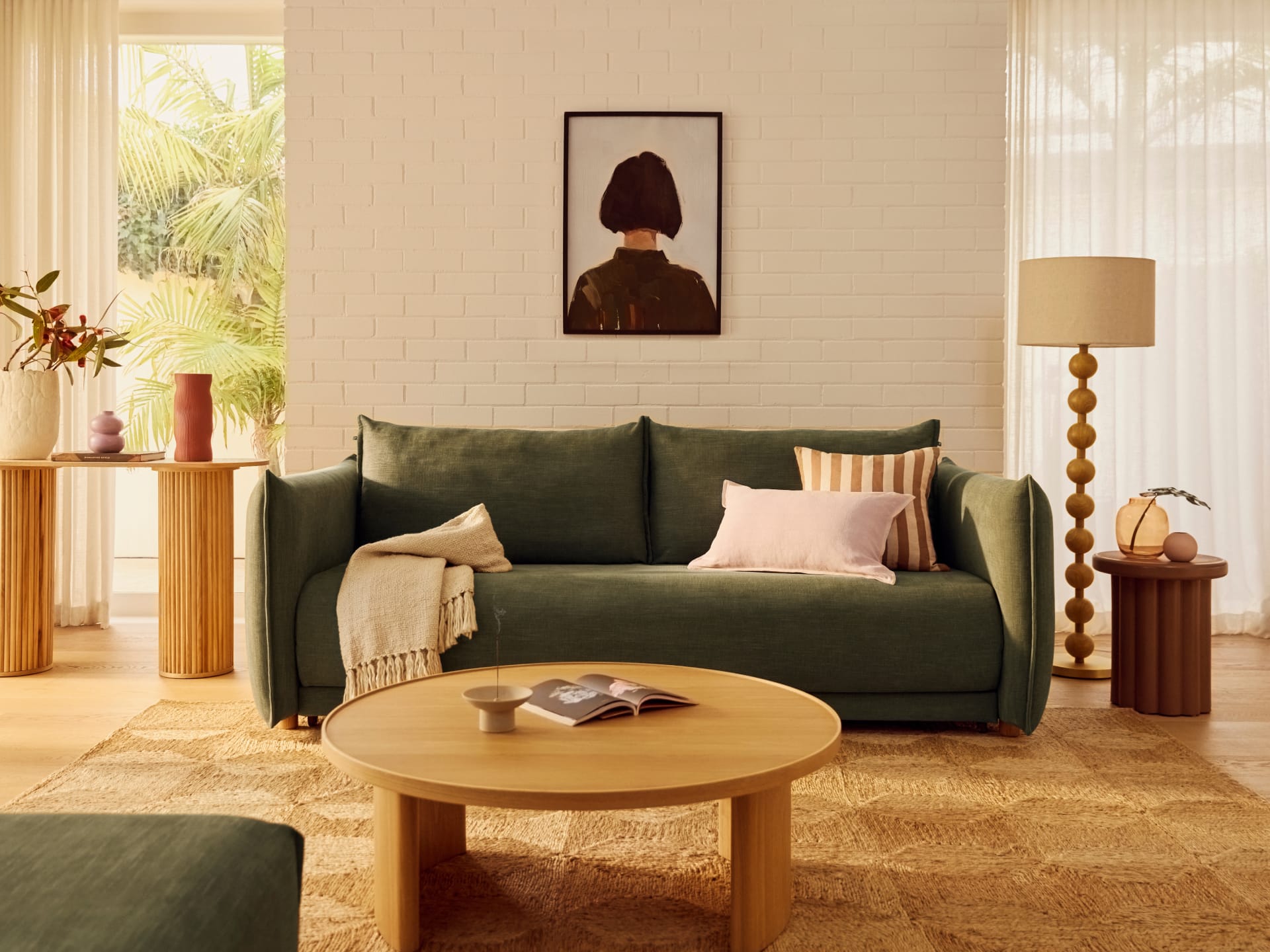 Koala Byron Sofa Bed [3rd Gen] in Forest Walk. Dark olive green two-seater couch in room with white brick wall, white sheers, round timber coffee table, and patterned jute floor rug. 