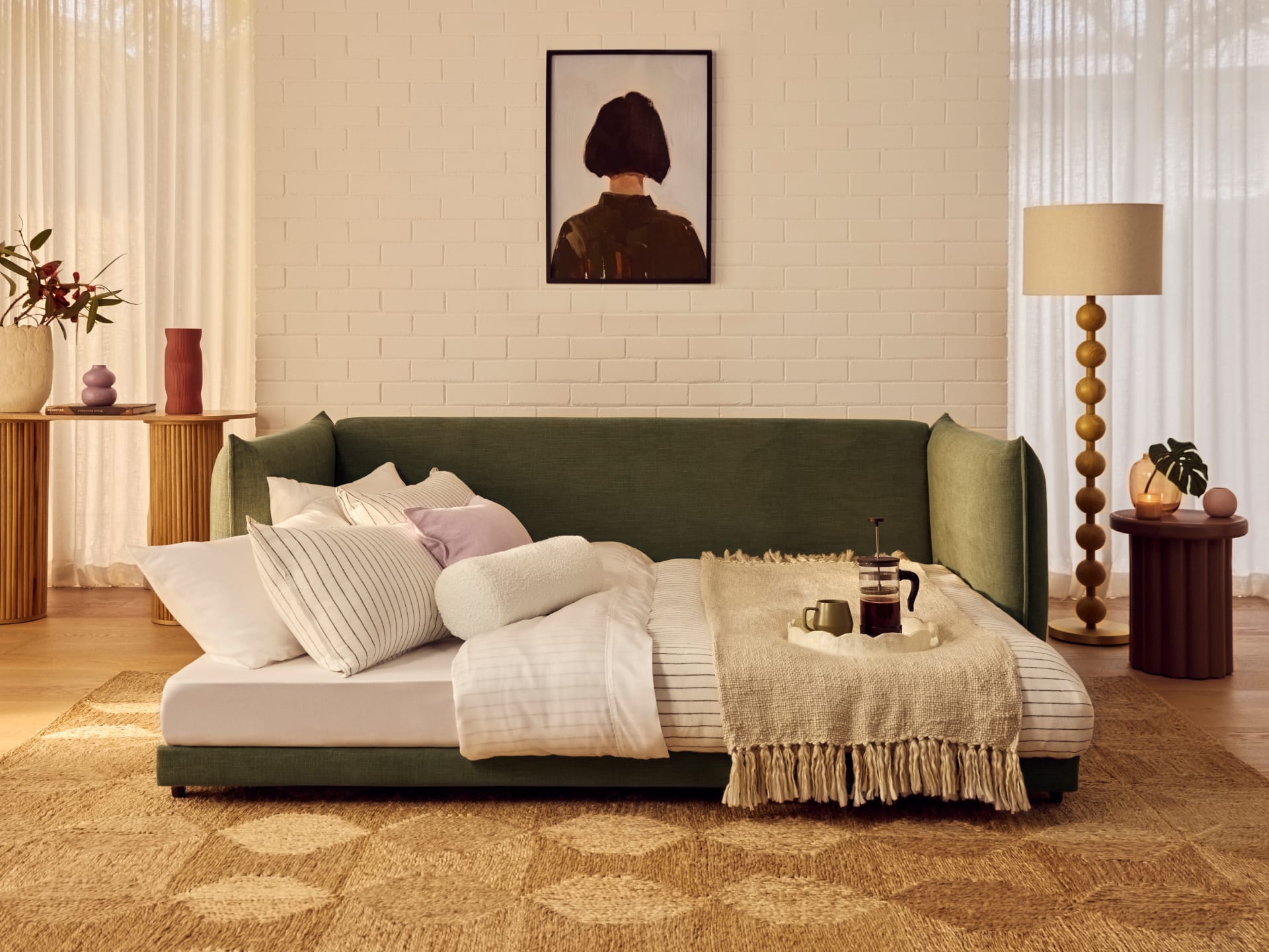 Koala Byron Sofa Bed [3rd Gen] in Forest Walk. Fold out sofa bed in dark olive-green fabric, with striped bedding and beige throw rug, in room with white brick walls. 