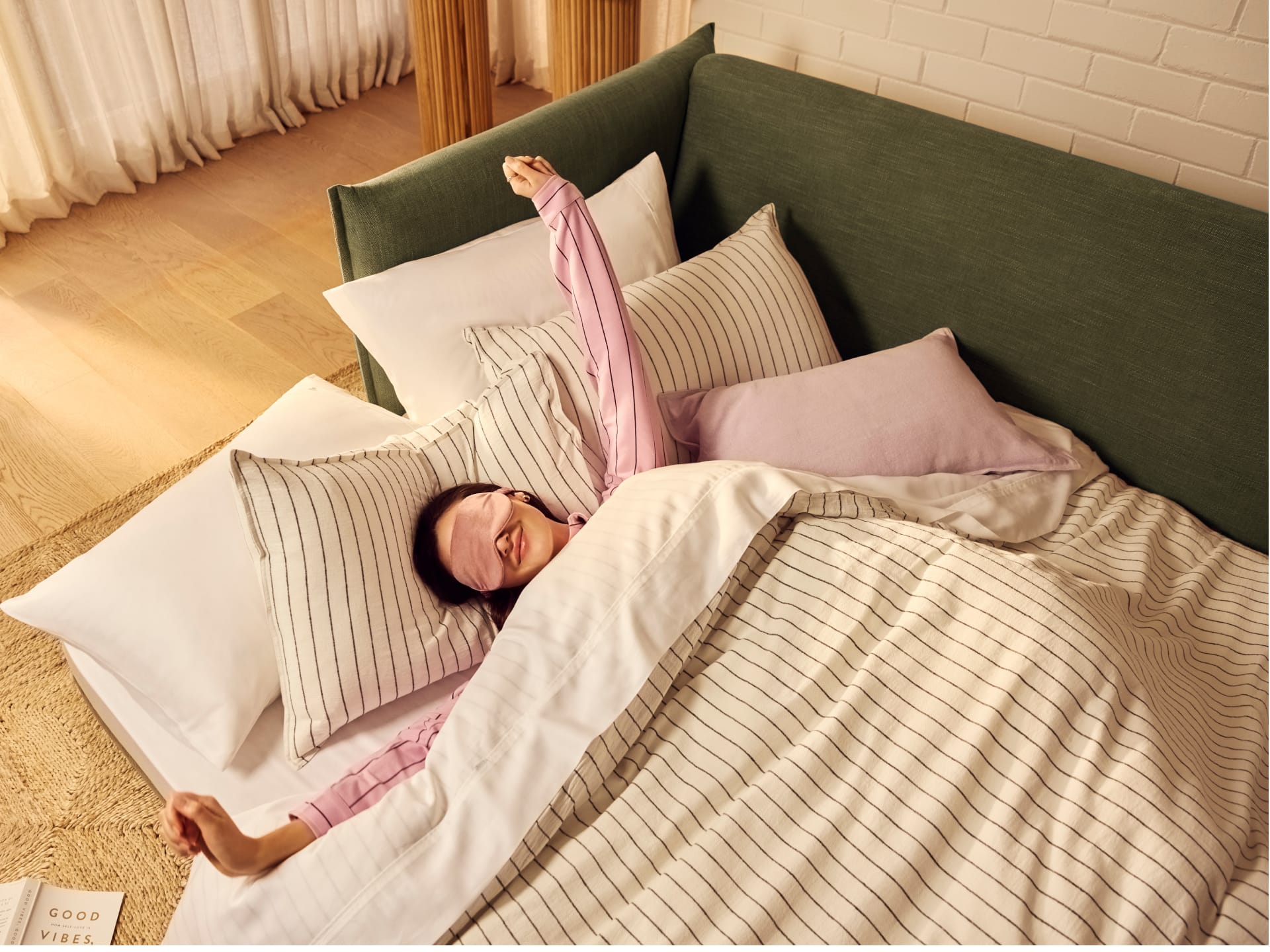 Koala Byron Sofa Bed [3rd Gen] in Forest Walk. Woman with pink eye mark and pink striped pyjamaes stretching, tucked into sofa bed with striped sheets, dark olive fabric finish and in a room with timber floors. 