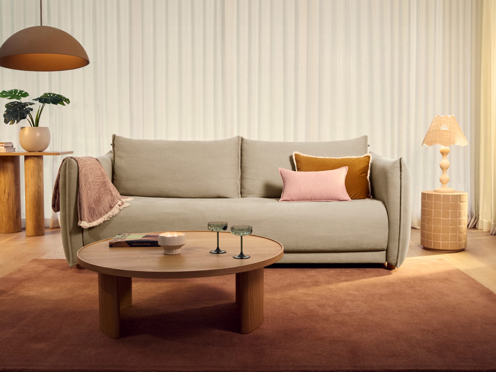 Koala Byron Sofa Bed [3rd Gen] in Lunar Glow. Grey two-seater couch with mustard and pink pillow, on terracotta floor rug, in front of white sheers, with round timber coffee table. 