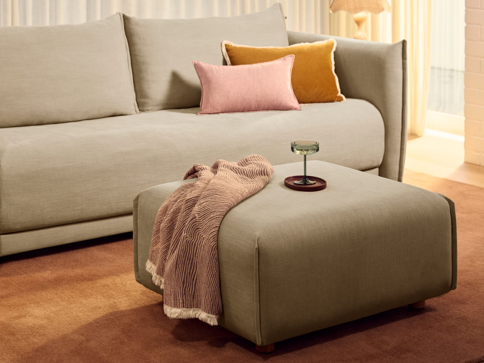 Koala Byron Sofa Bed [3rd Gen] in Lunar Glow. Grey two-seater lounge with matching grey fabric ottoman, on top of terracotta rug, with pink and mustard cushions on couch. 