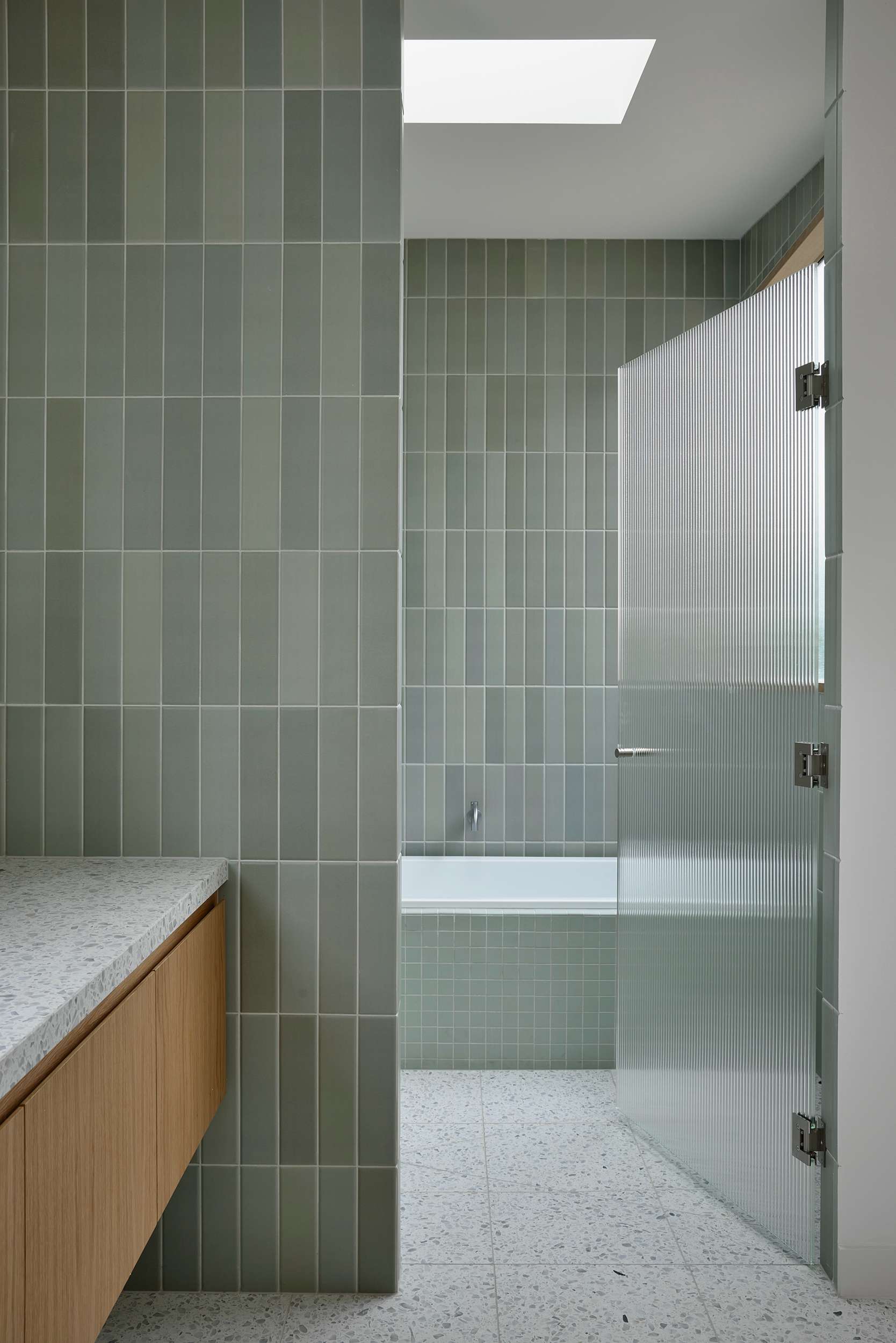 Home Pavilion by MRTN Architects. Bathroom with white terrazzo flooring, green wall tiles, timber cabinetry and fluted glass door. 