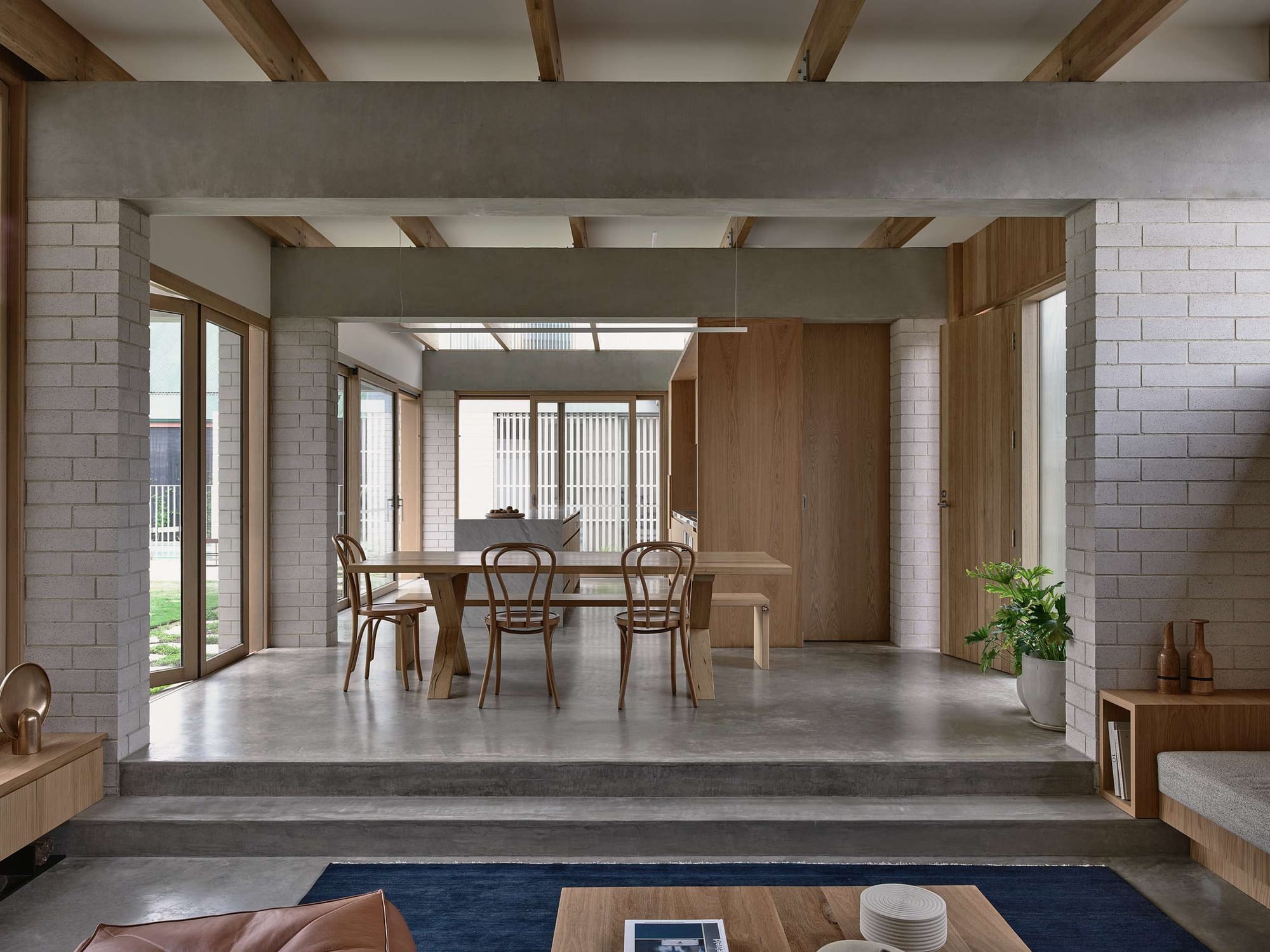 Home Pavilion by MRTN Architects. Timber dining table and chairs on elevated concrete surface, with exposed concrete beams overhead and white brick pillars. 