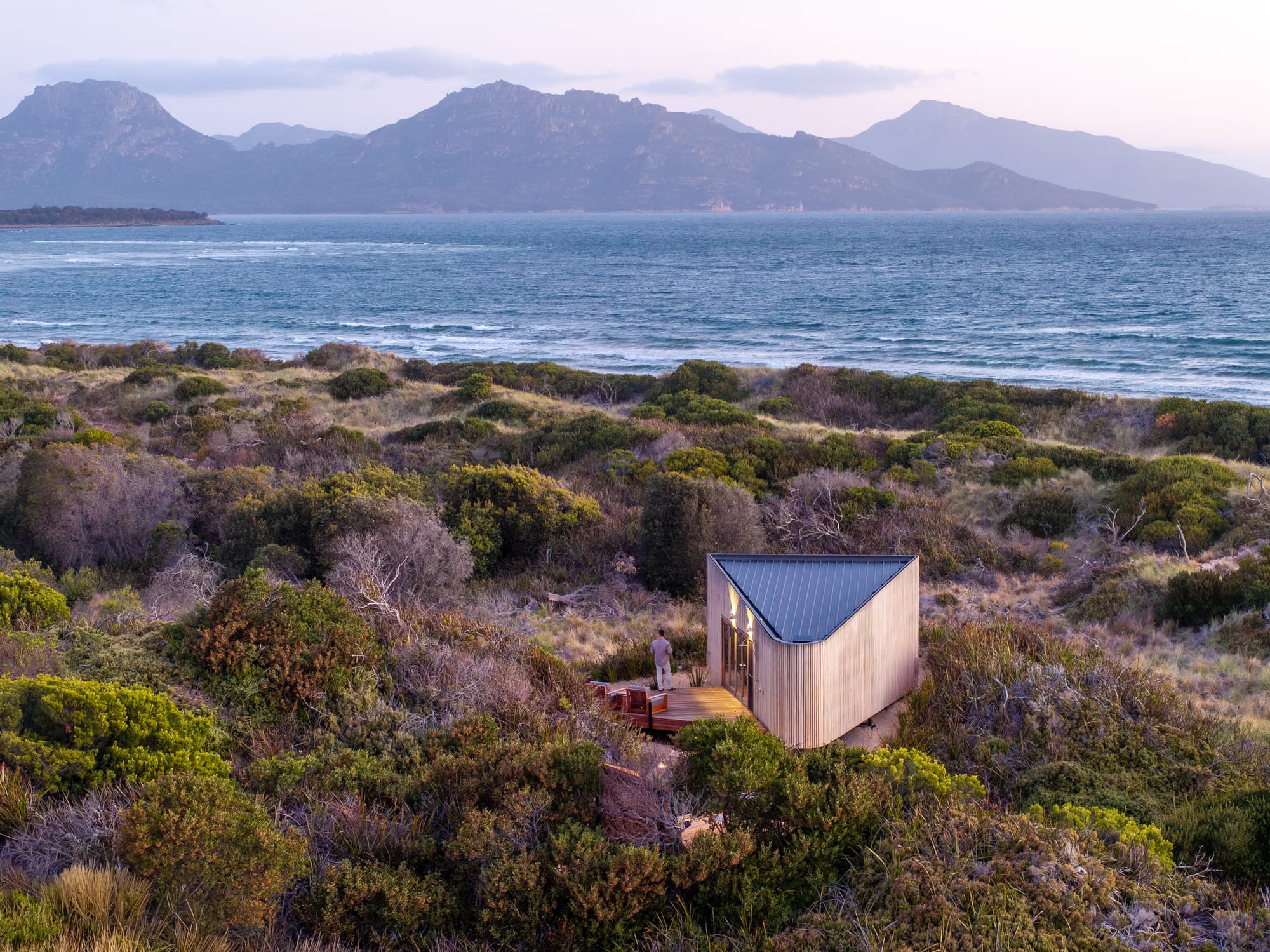 A landscape shot of Studio Tasmania located in the coastal bush with a man standing on the deck looking out to the ocean.