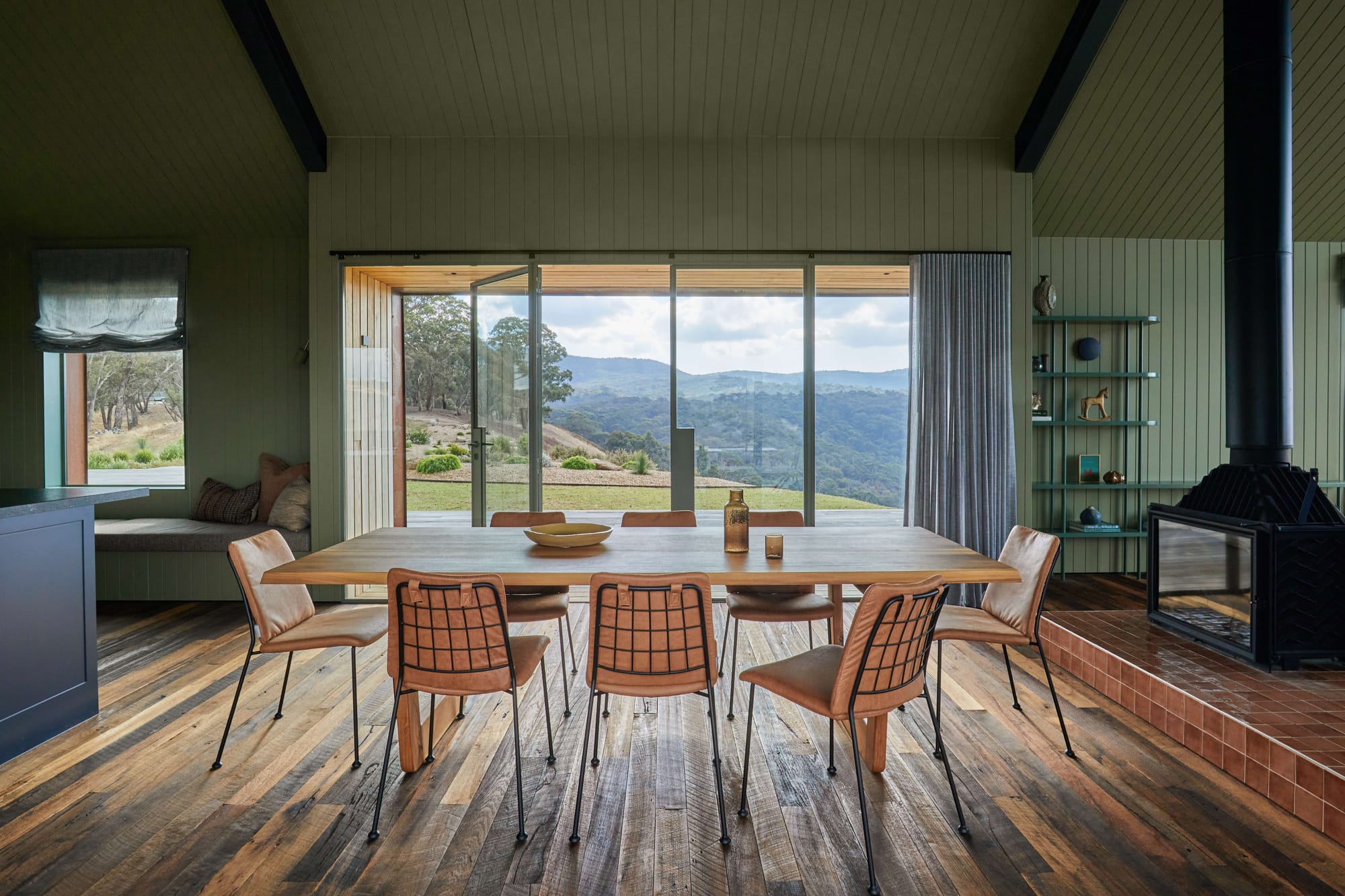 Horan's Track by TANDEM Design Studio. Photography by Stephanie Rooney. Interior with timber floors, clad walls painted sage green, windows overlooking rolling landscape, and simple timber dining table. 