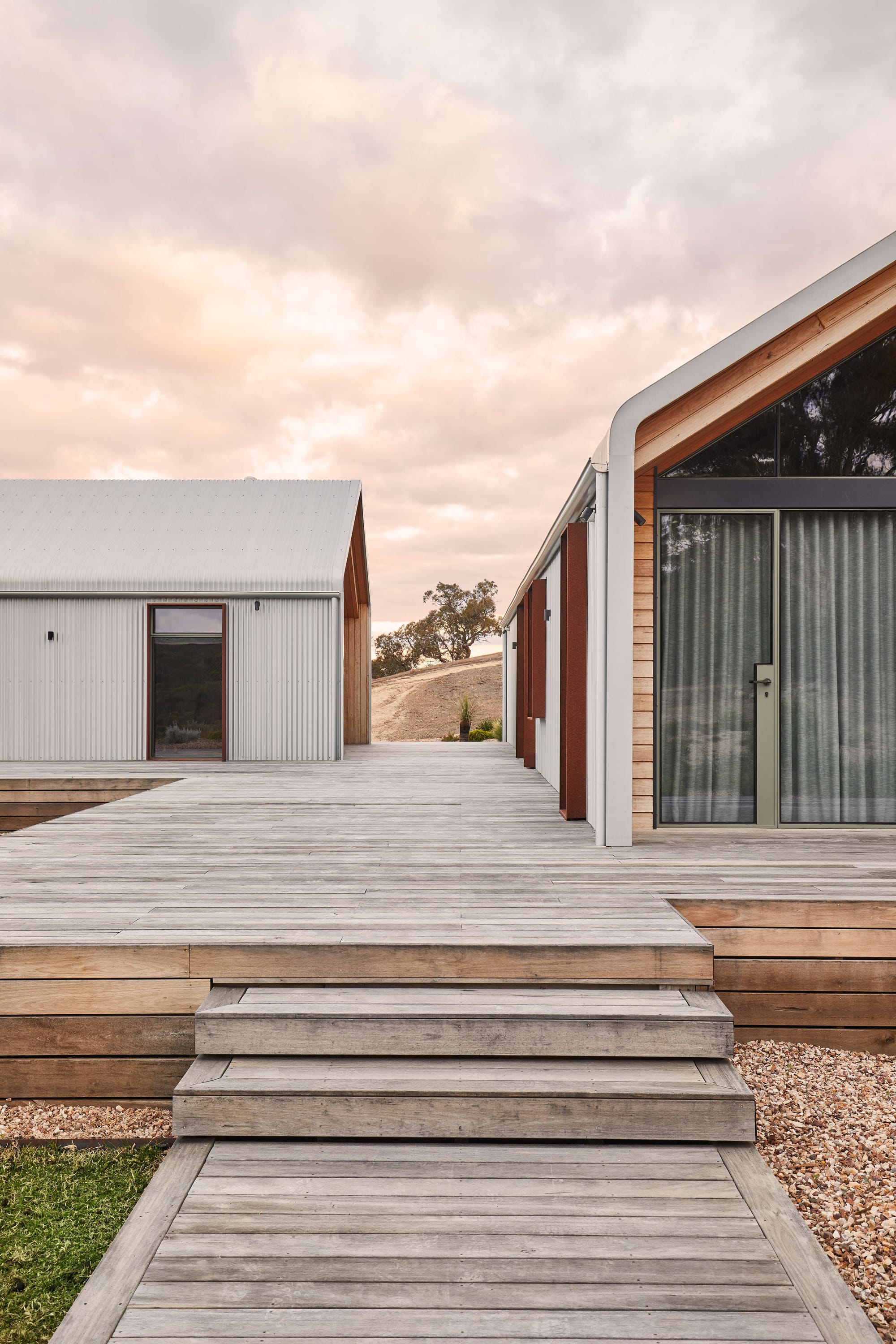 Horan's Track by TANDEM Design Studio. Photography by Stephanie Rooney. Two pavilion style structures connected by weathered timber decking, with timber stairs in foreground and rural setting in background. 