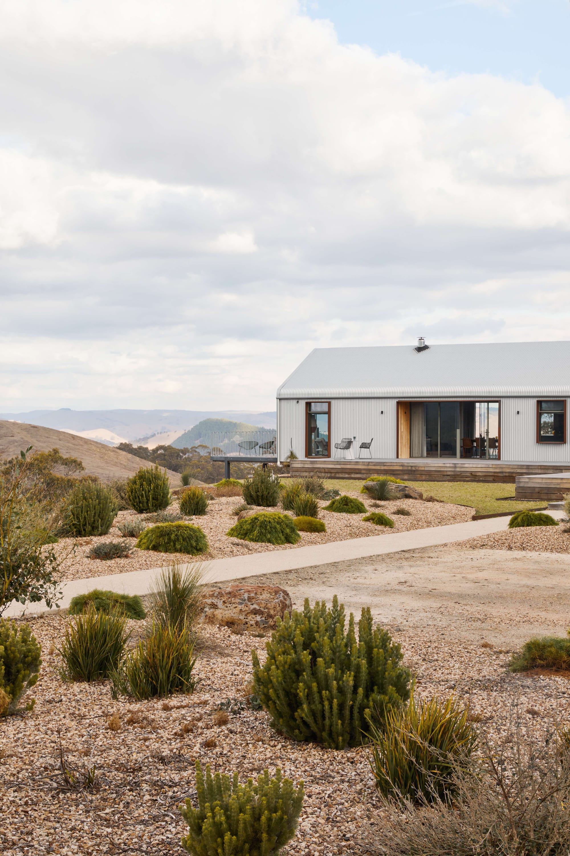 Horan's Track by TANDEM Design Studio. Photography by Stephanie Rooney. Aluminum clad paviion style home with timber deck and balcony extending over cliff face, with simple native garden in foreground and rolling hills in background. 