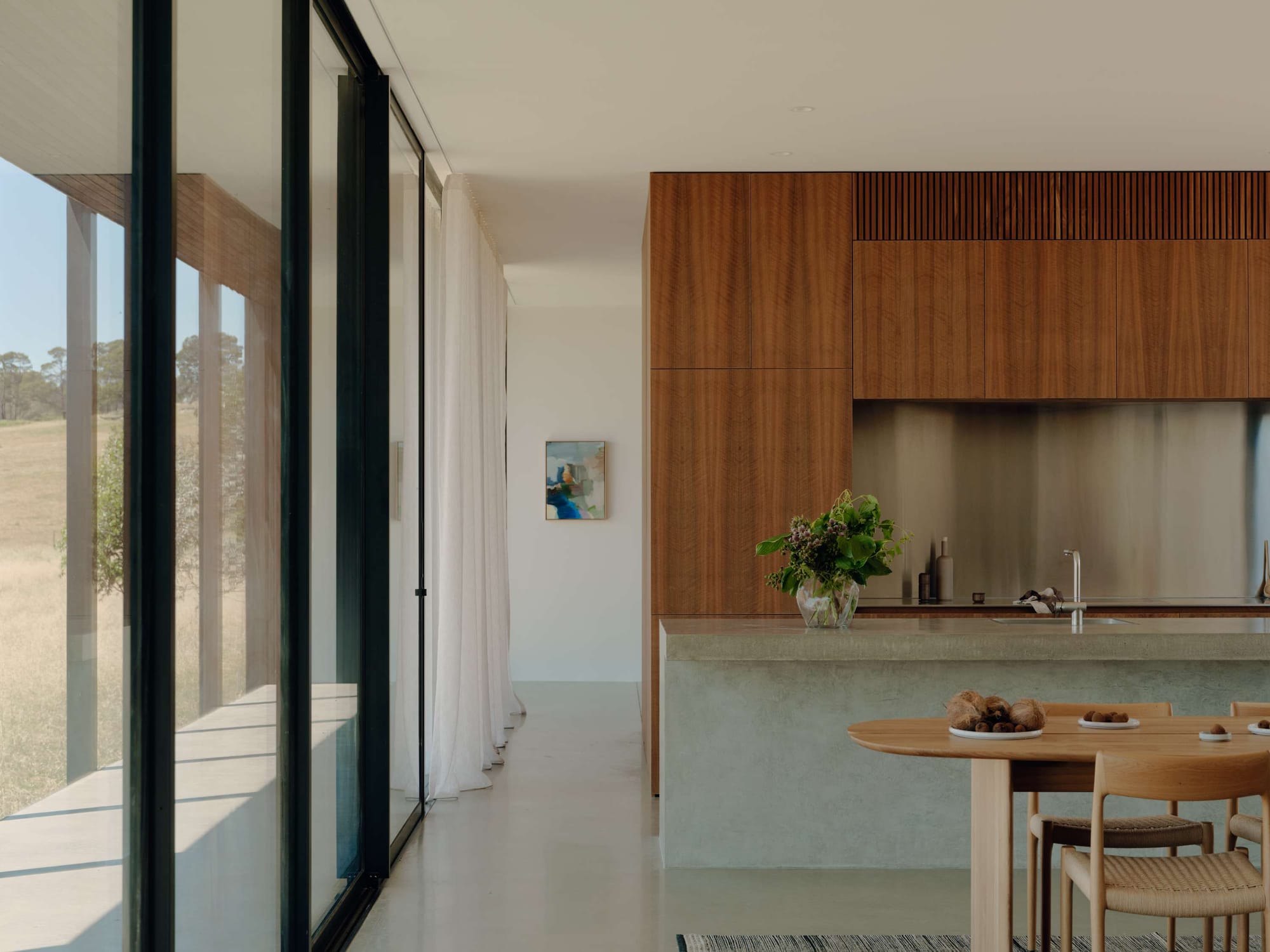 Marraweeney by Tom Robertson Architecture. Photography by Tom Ross. Kitchen and dining space in home with polished concrete floors, full height glass windows, timber kitchen cabinetry and dining table. 