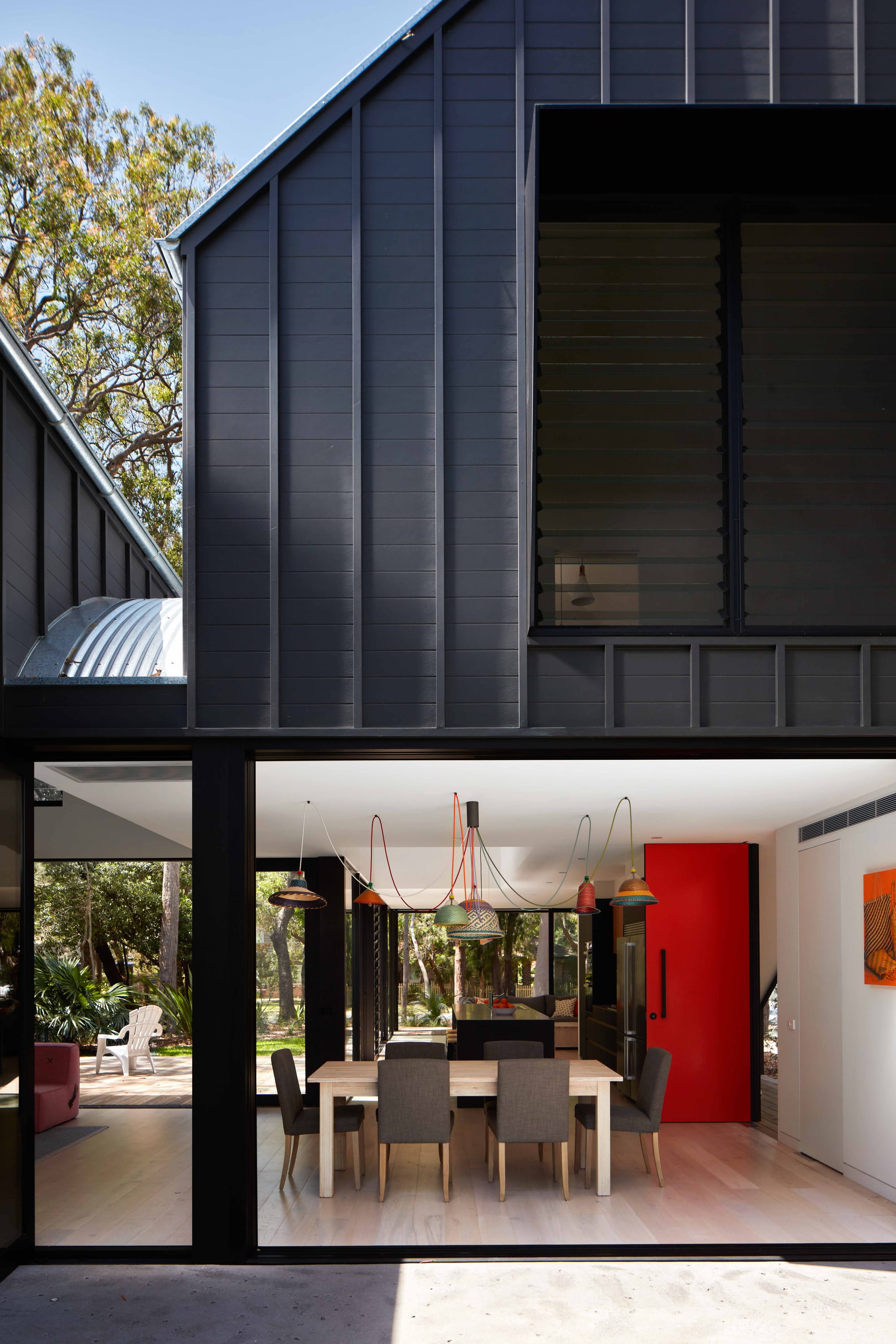 Pearl Beach II by COSO Architecture. Dinning are with large windows. Wooden dinning table with grey chairs. Red door and colourful lamps.