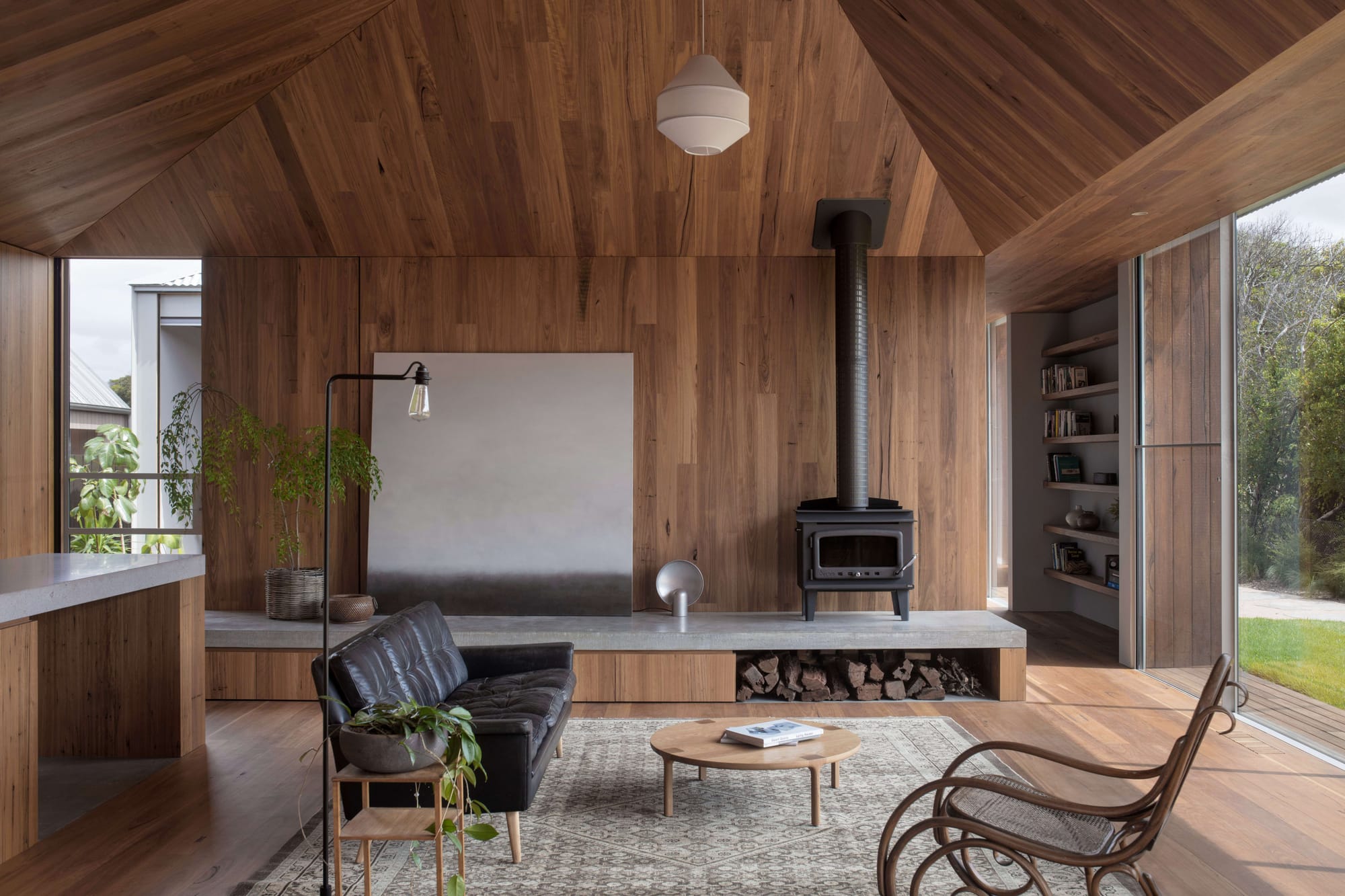Point Lonsdale House by Edition Office. Photographed by Ben Hosking. Living are with wooden walls and wooden floors. Wooden ceiling and indoor fireplace. Raised platform and black leather couch.