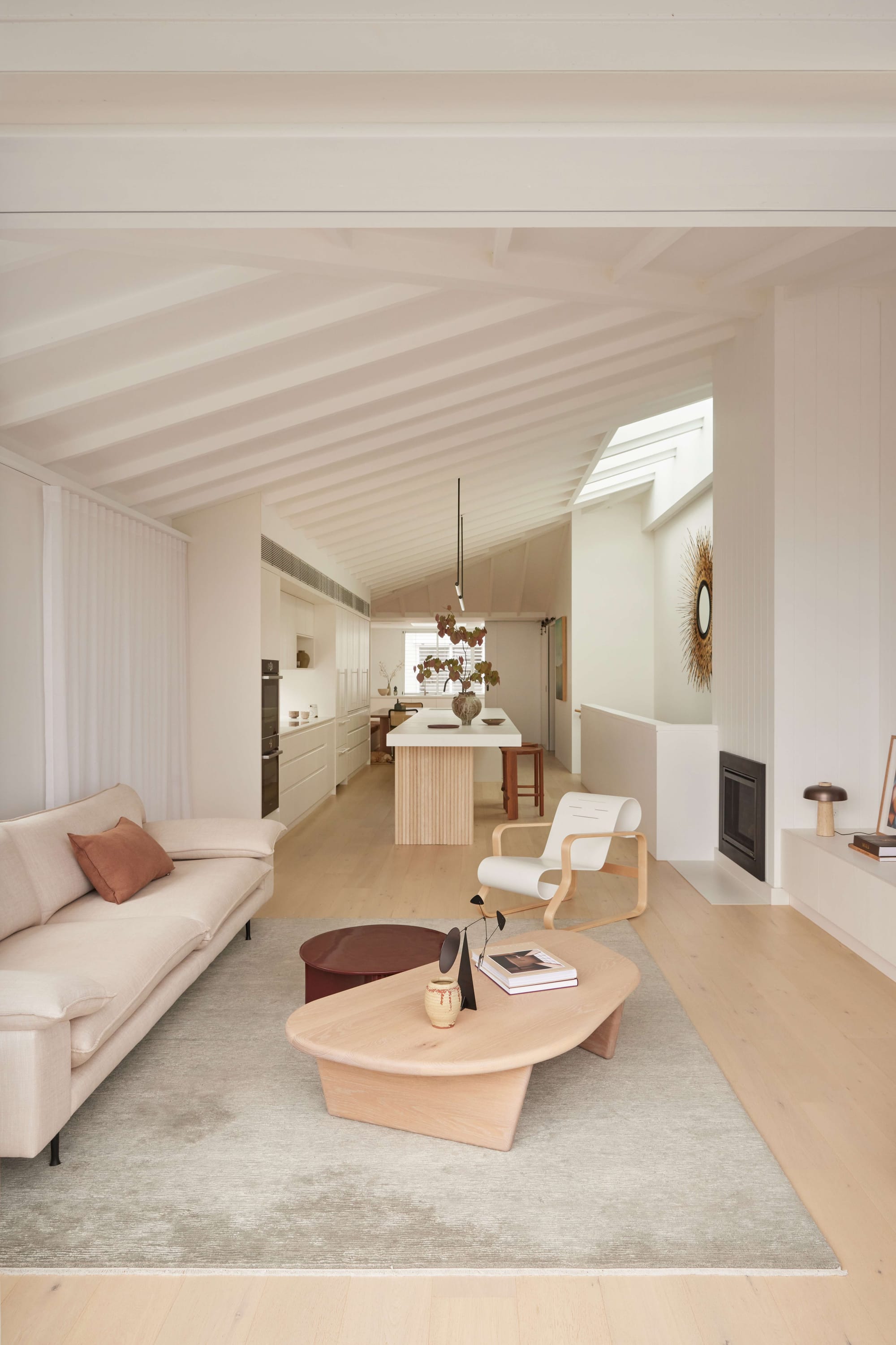 Seaside House by Carla Middleton Architecture. Photographed by Alana Landberry. Living area with wooden floors and beige couch. Kitchen with white cabinets and slanted ceiling.