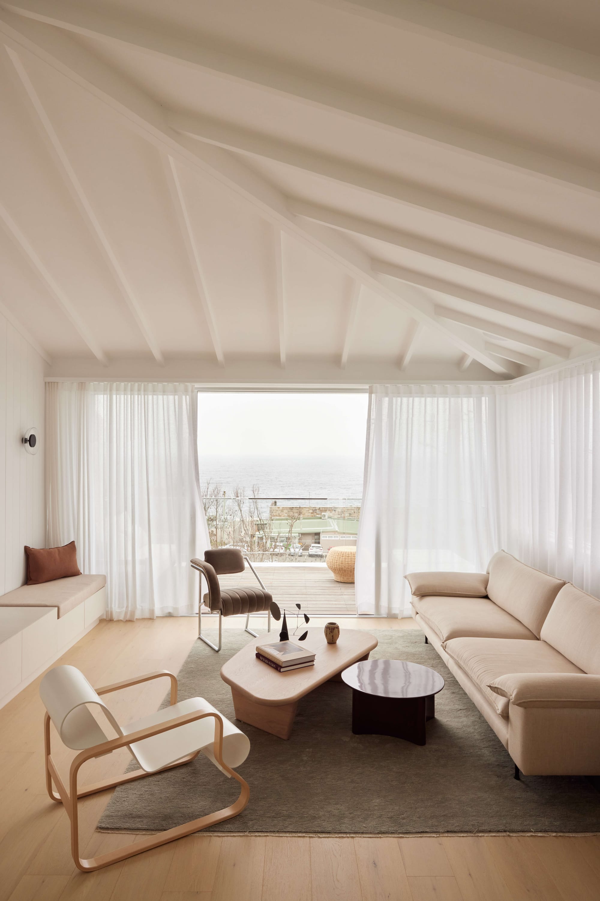 Seaside House by Carla Middleton Architecture. Photographed by Alana Landberry. Living area with balcony. View of the beach. Wooden coffe table and beige couch. White walls with sheer white curtains and wooden floors.