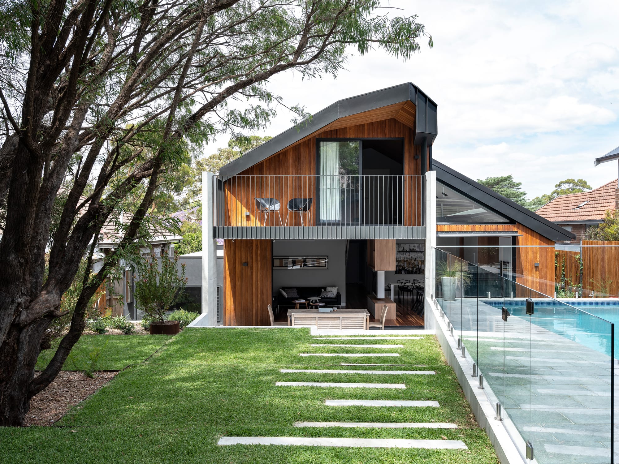Contemporary house with sloped rooflines, timber cladding, balcony, and open ground floor overlooking a lawn and swimming pool.