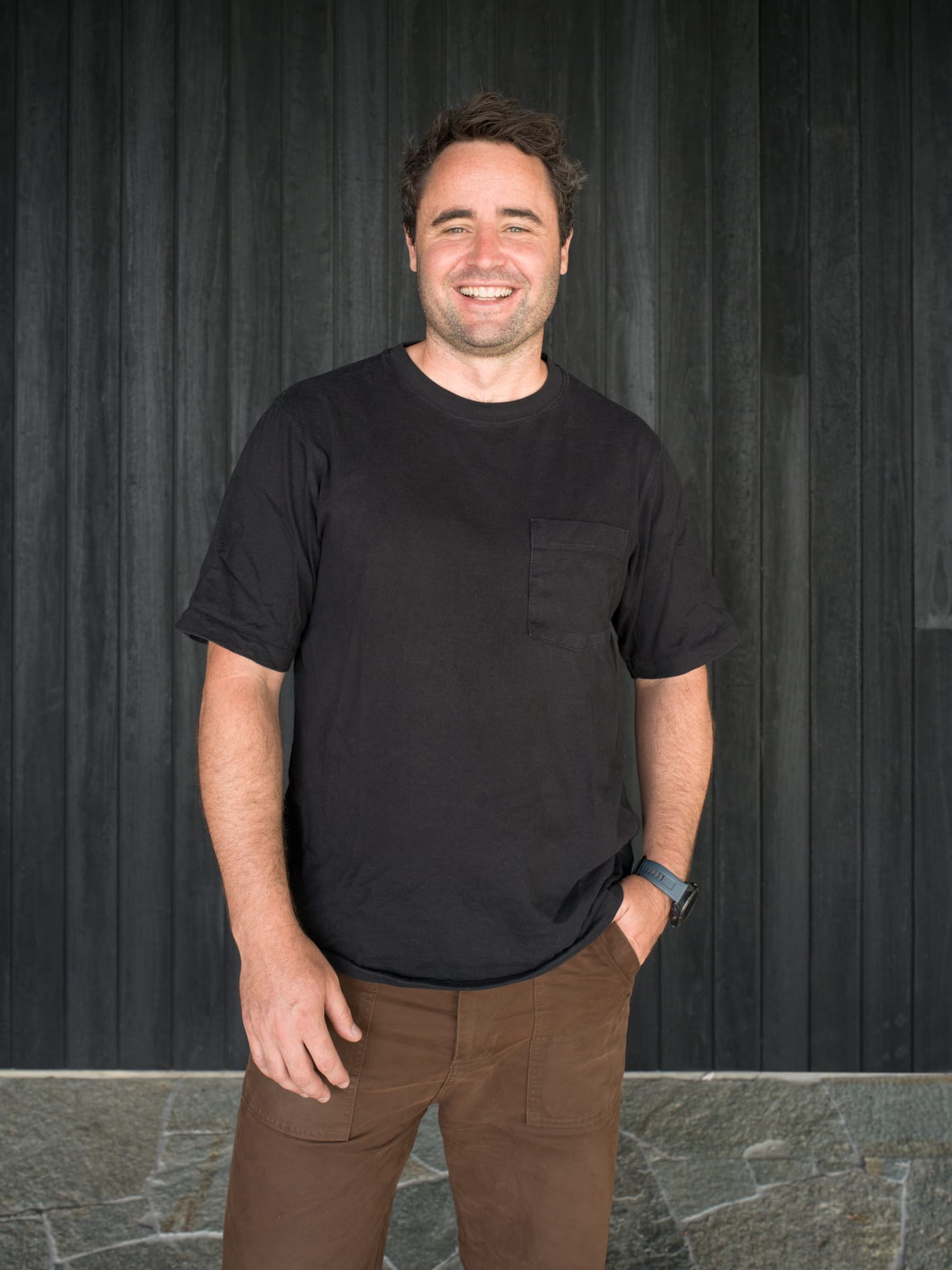 Portrait of a man standing against a dark timber wall, wearing a black t-shirt and brown pants, smiling at the camera.