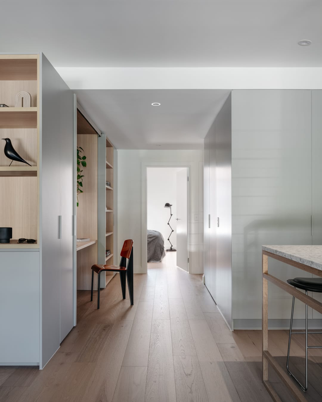 Minimal interior corridor with light timber flooring, built-in joinery, and a view through to a softly lit bedroom.