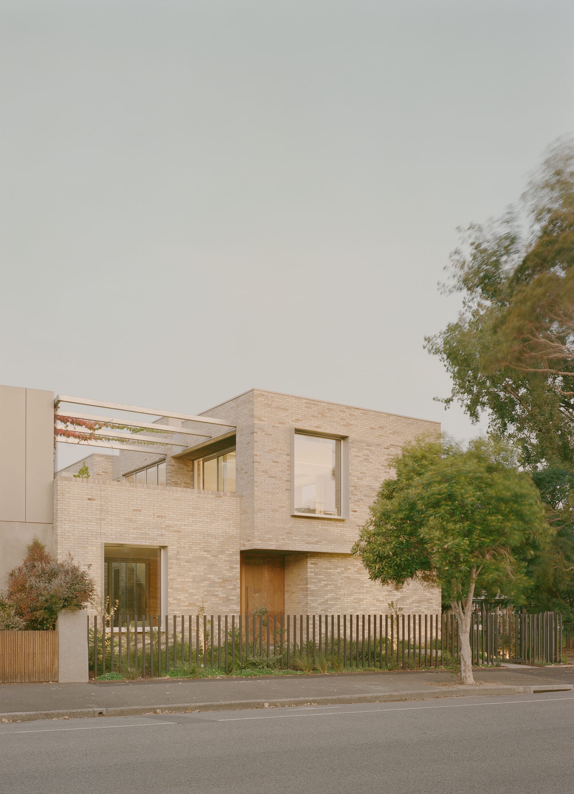 Two-storey brick house with a minimalist facade, large windows, and landscaped front garden along a suburban street.