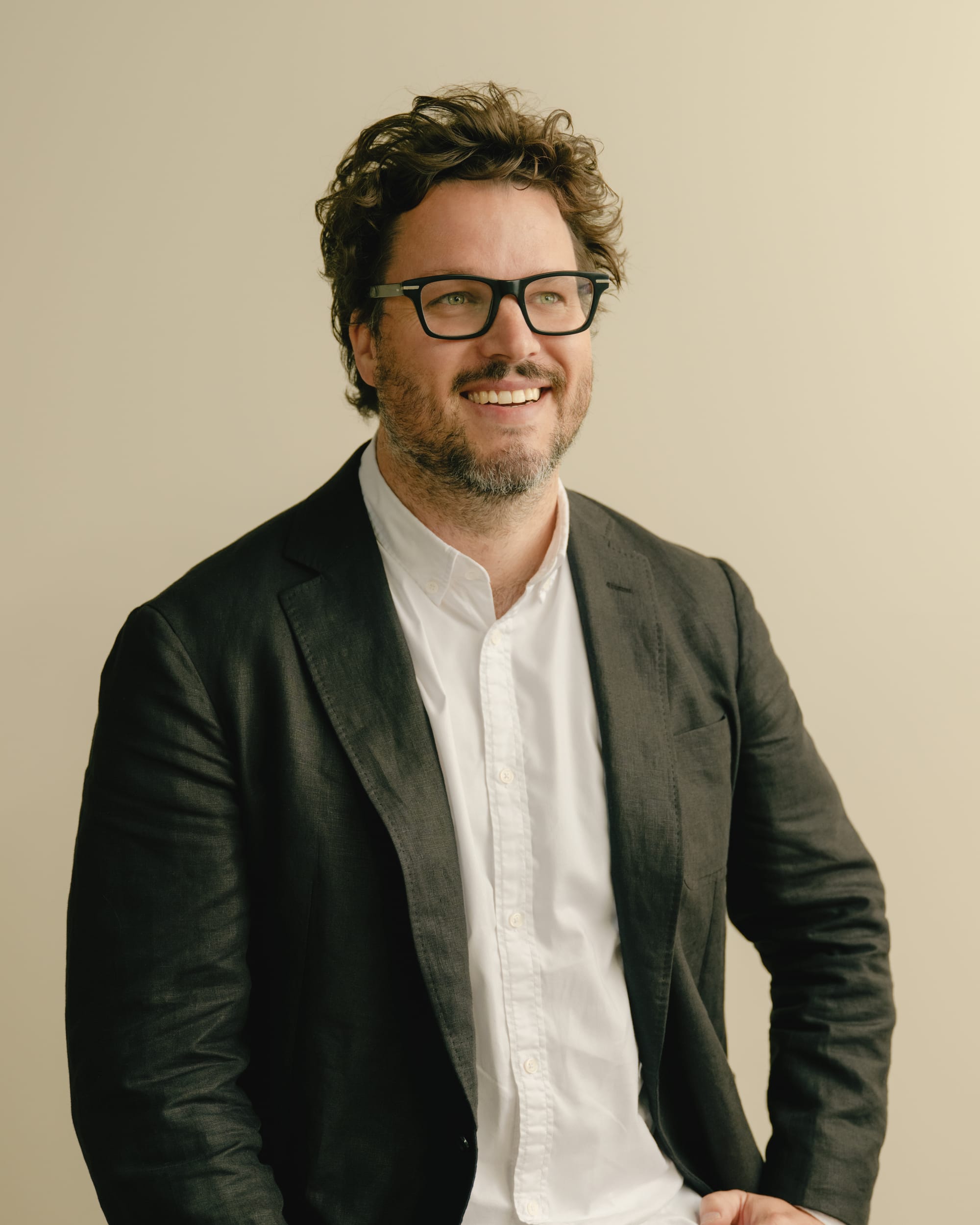 Portrait of a man wearing glasses and a black blazer, seated against a neutral background and smiling.