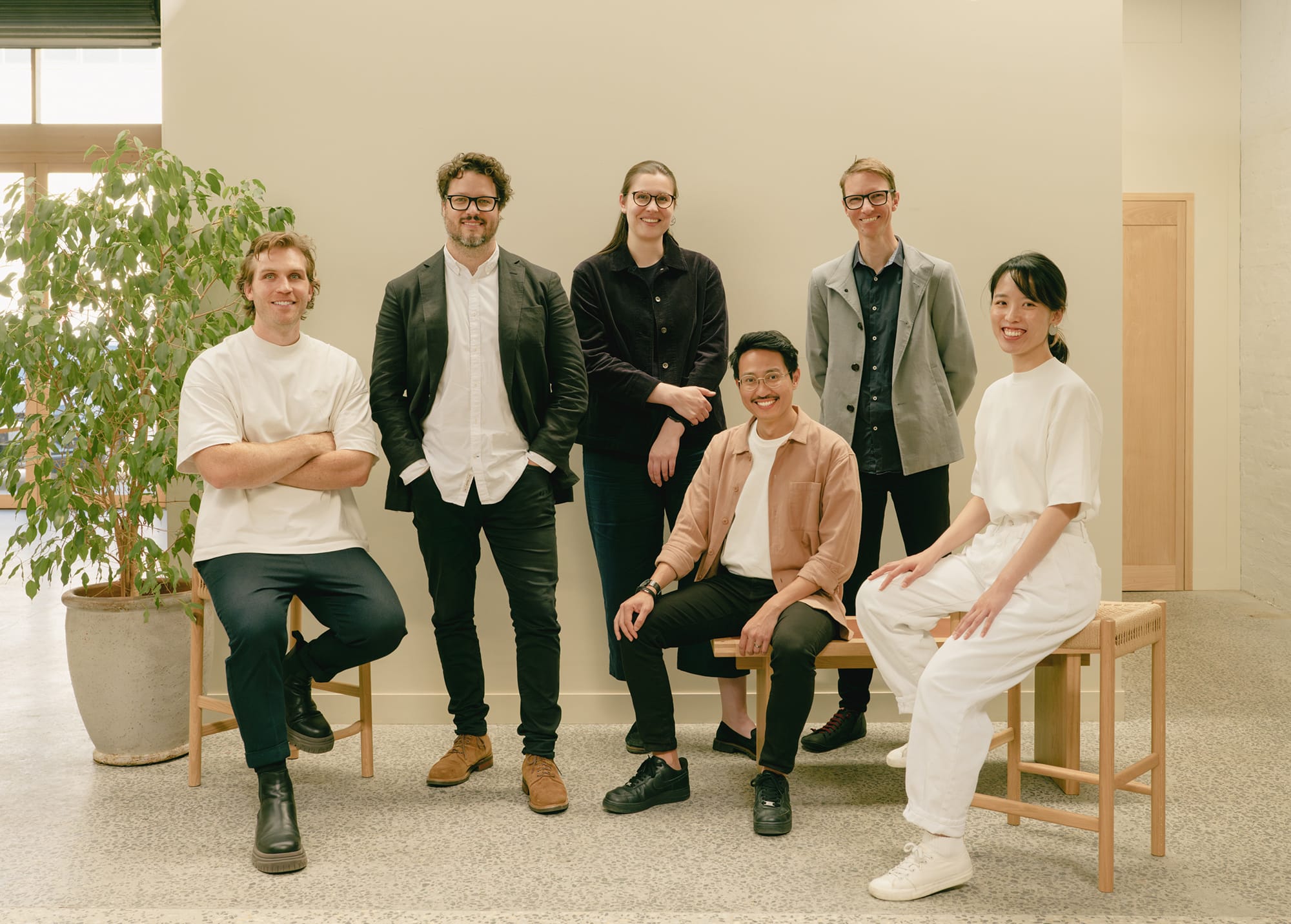Group portrait of six architects standing and seated in a studio space with a neutral interior and indoor plant.