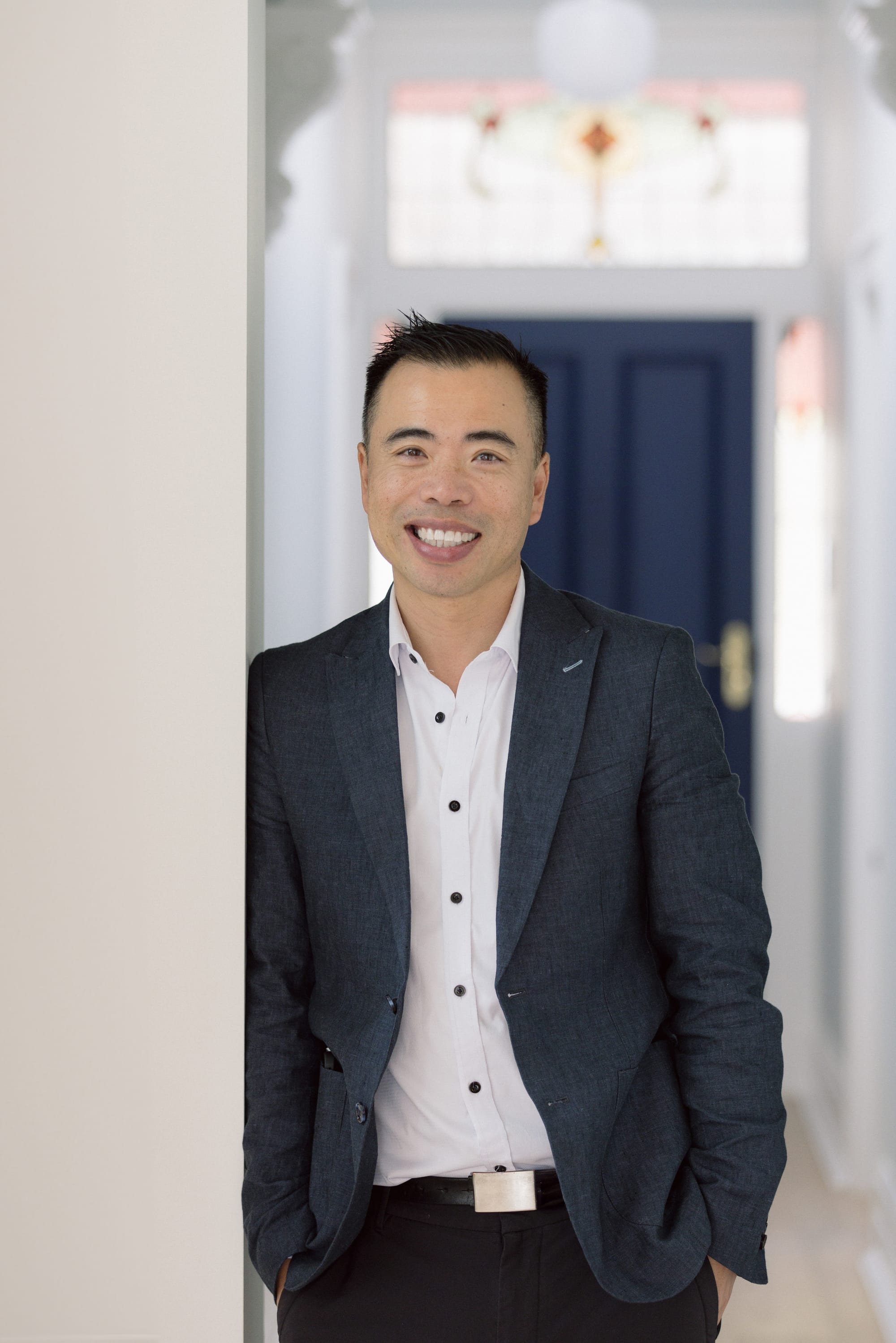 Portrait of a man in a blazer and white shirt, smiling while standing in a softly lit interior hallway.