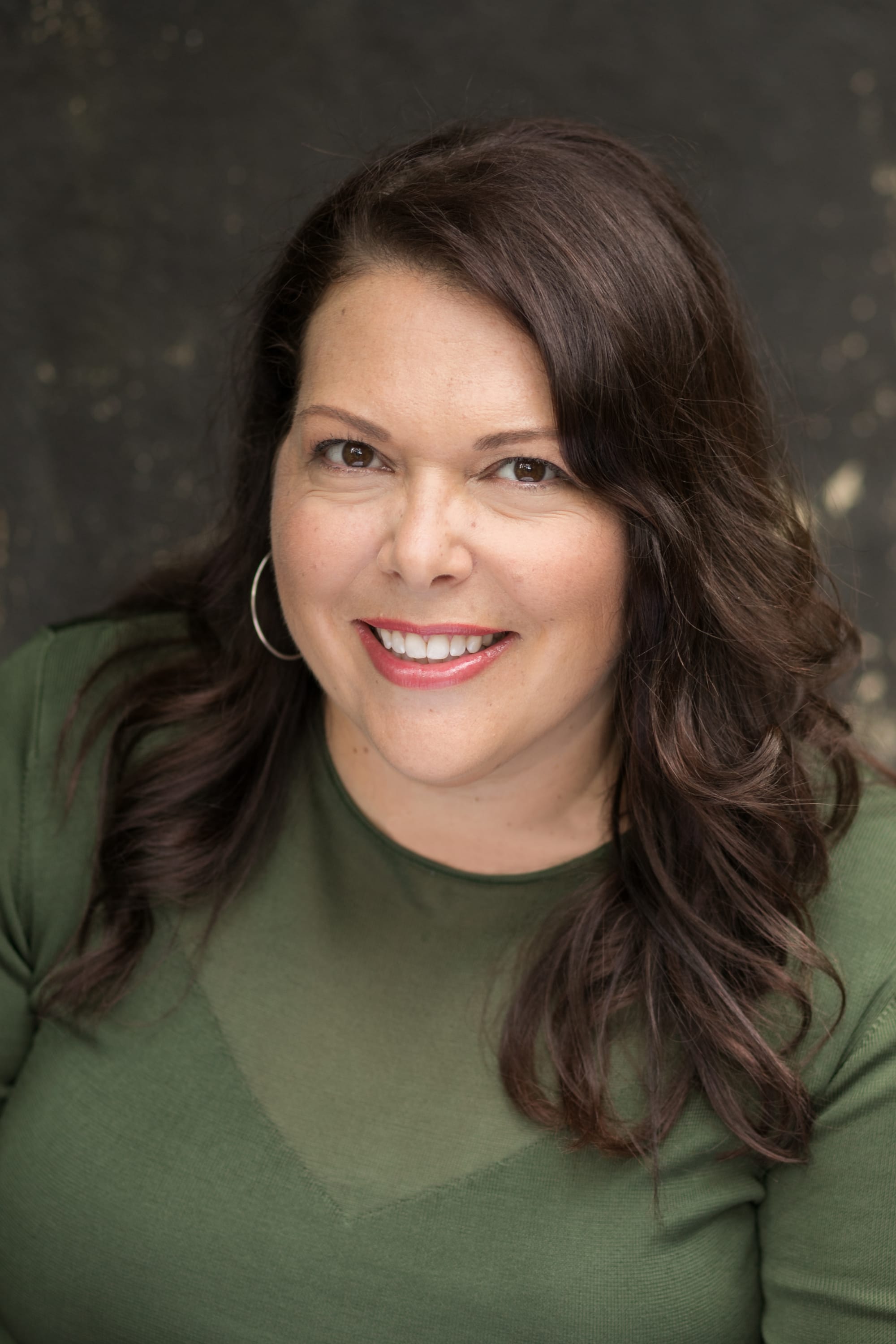 Smiling woman, Melonie Bayl-Smith, with shoulder-length dark hair wearing a green top, photographed against a softly blurred dark background.