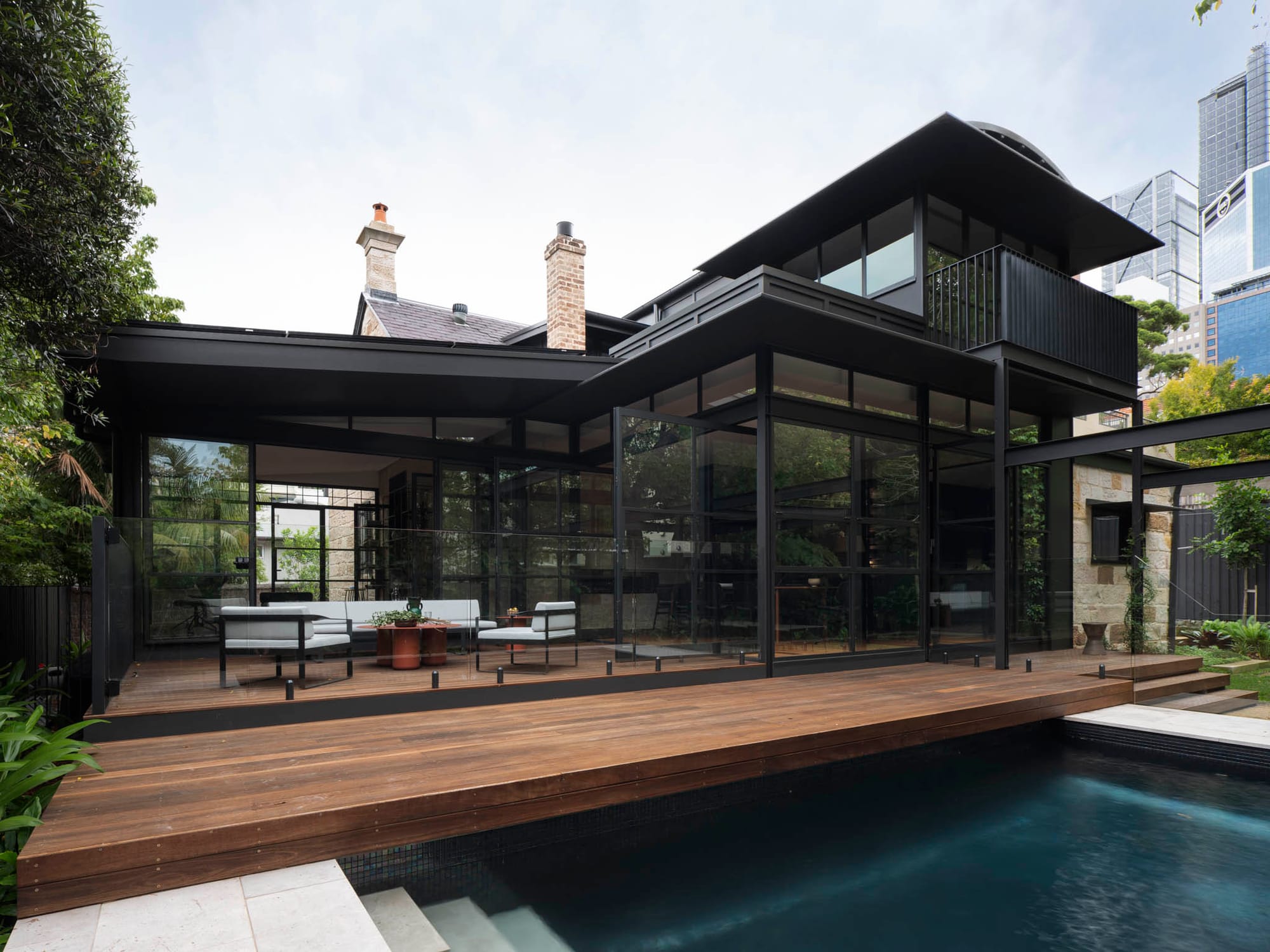 Modern black-clad home extension with floor-to-ceiling glazing, opening onto a timber deck beside a pool.