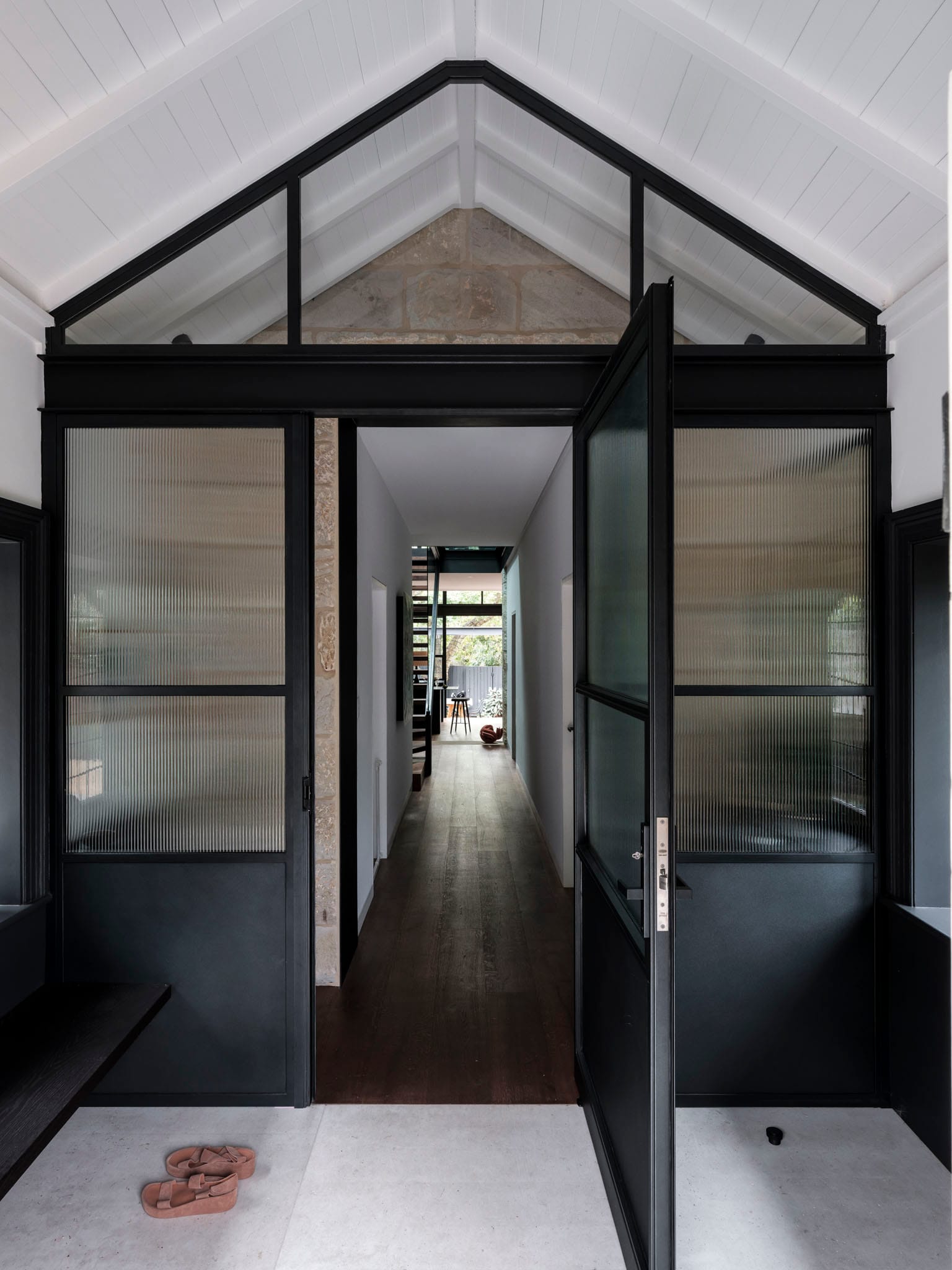 Symmetrical entry with white pitched ceiling, black-framed glazed doors, and a long hallway leading to a light-filled rear space.