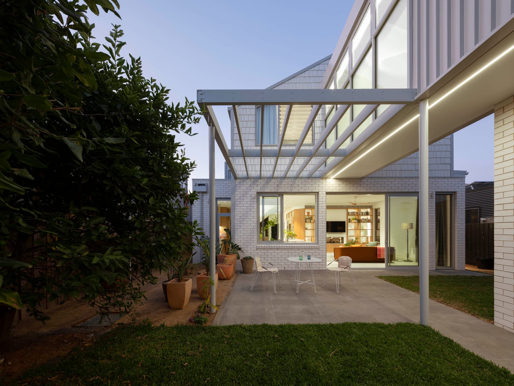 Light brick home with a steel pergola over a paved courtyard, warm interior lighting visible through large windows at dusk.