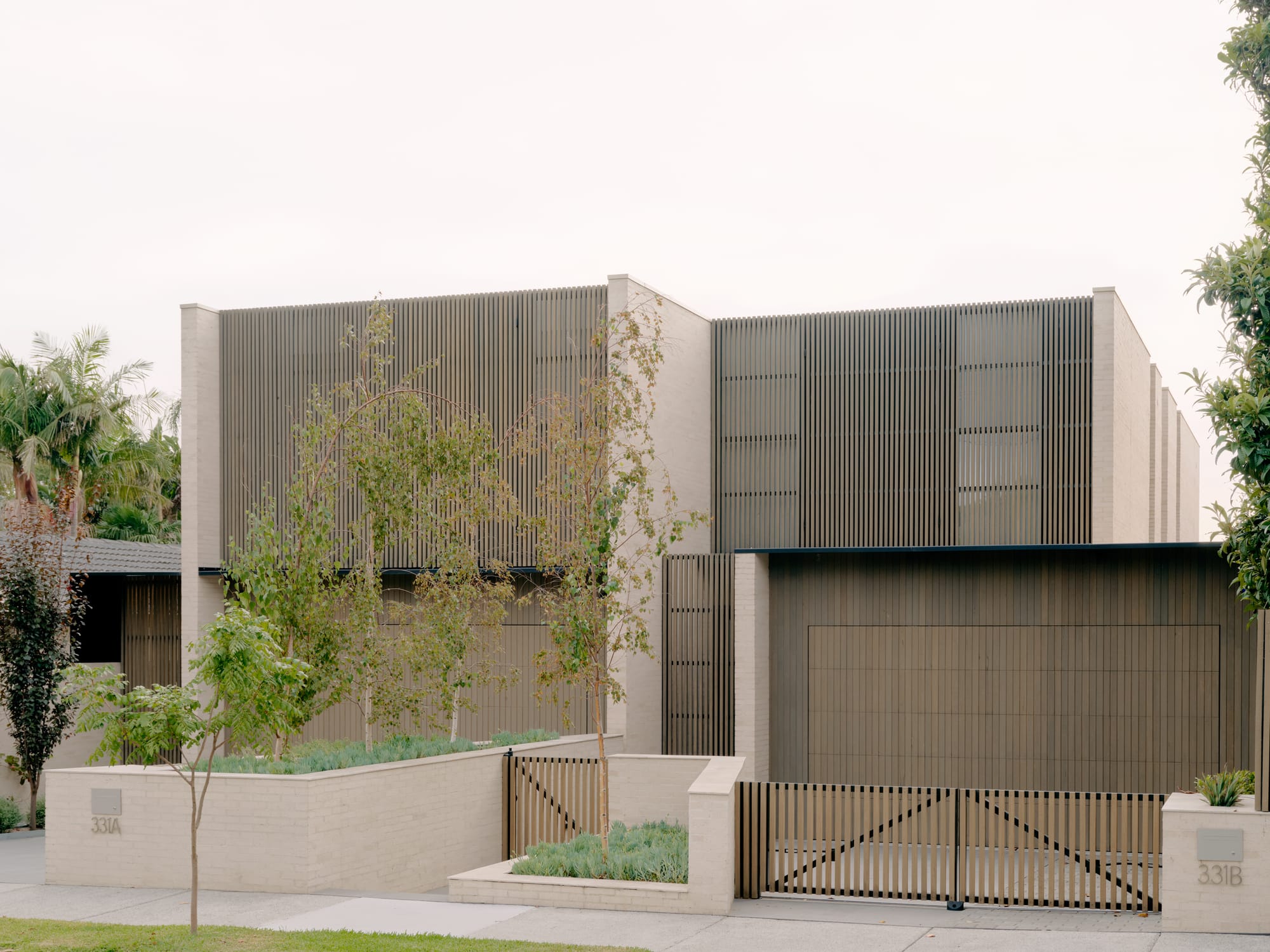 Contemporary residential facade with light brick, vertical screening, and a landscaped front garden.