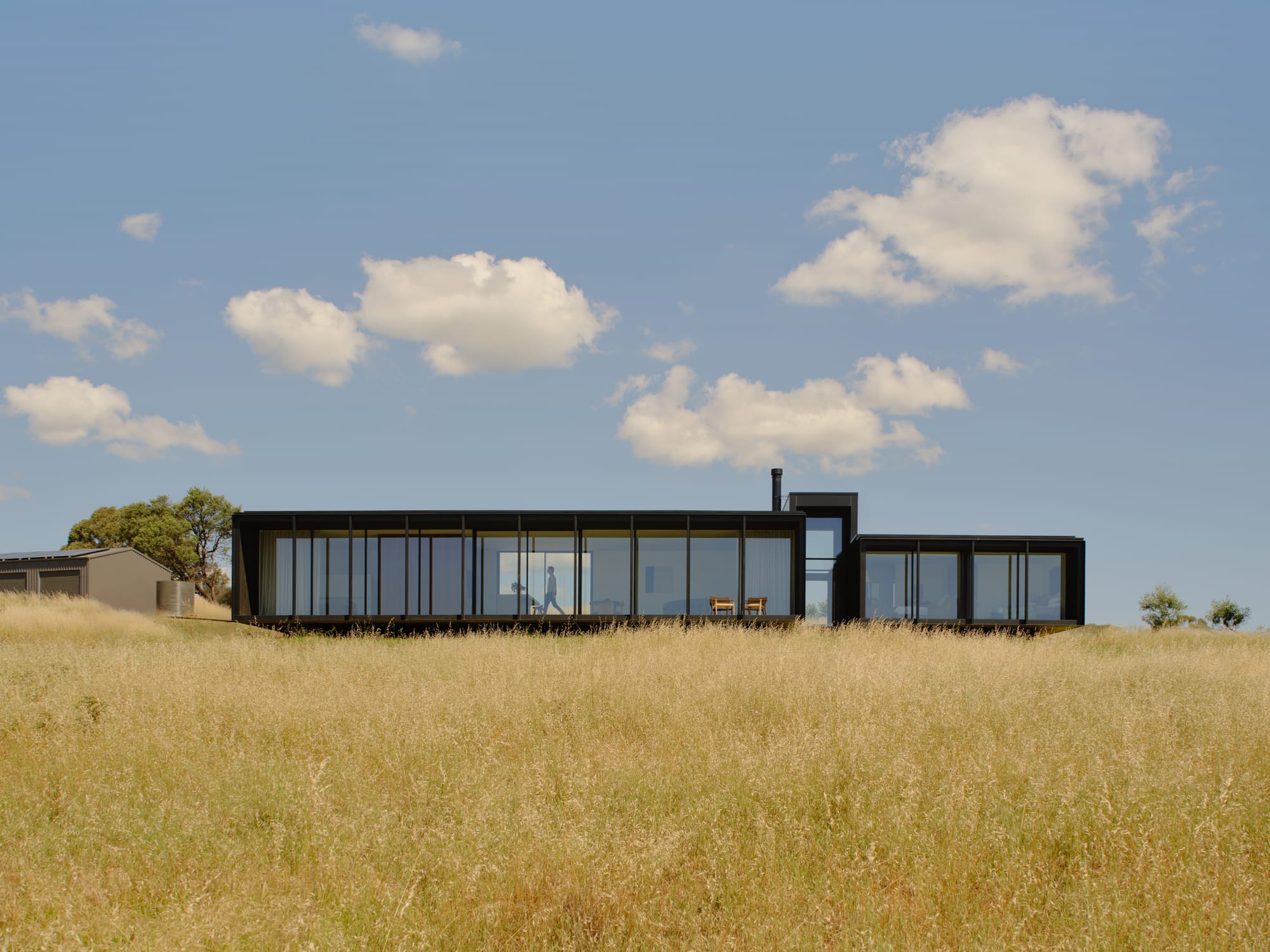 Low-profile modern house with dark cladding set within an open grassy landscape under a blue sky.