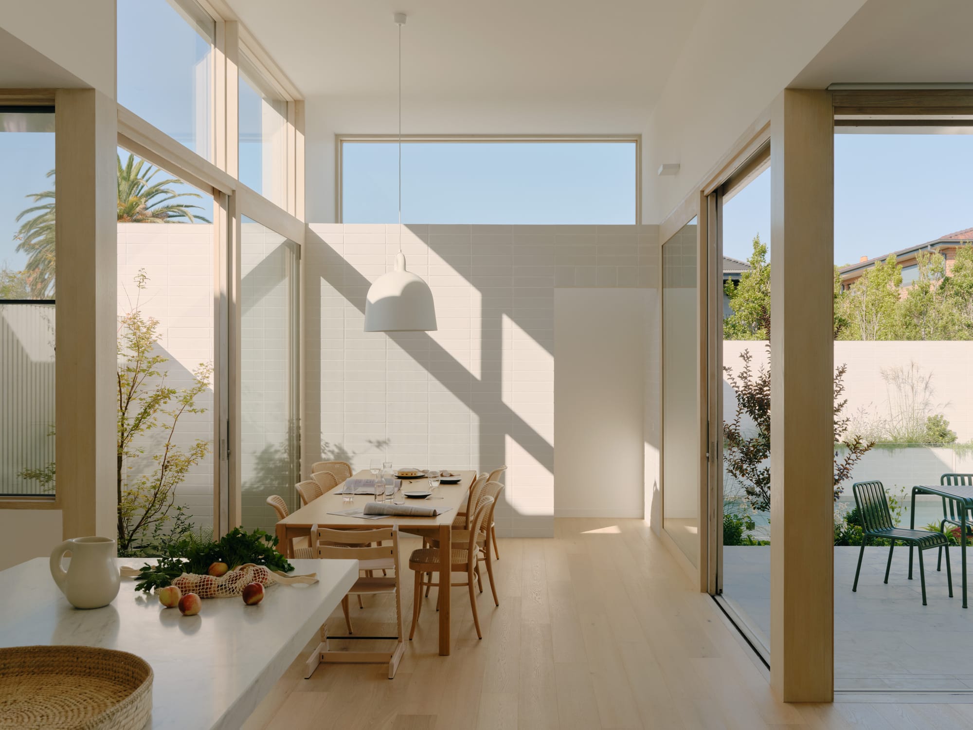 Light-filled dining space with timber furniture, large windows, and strong sunlight casting shadows across the interior.