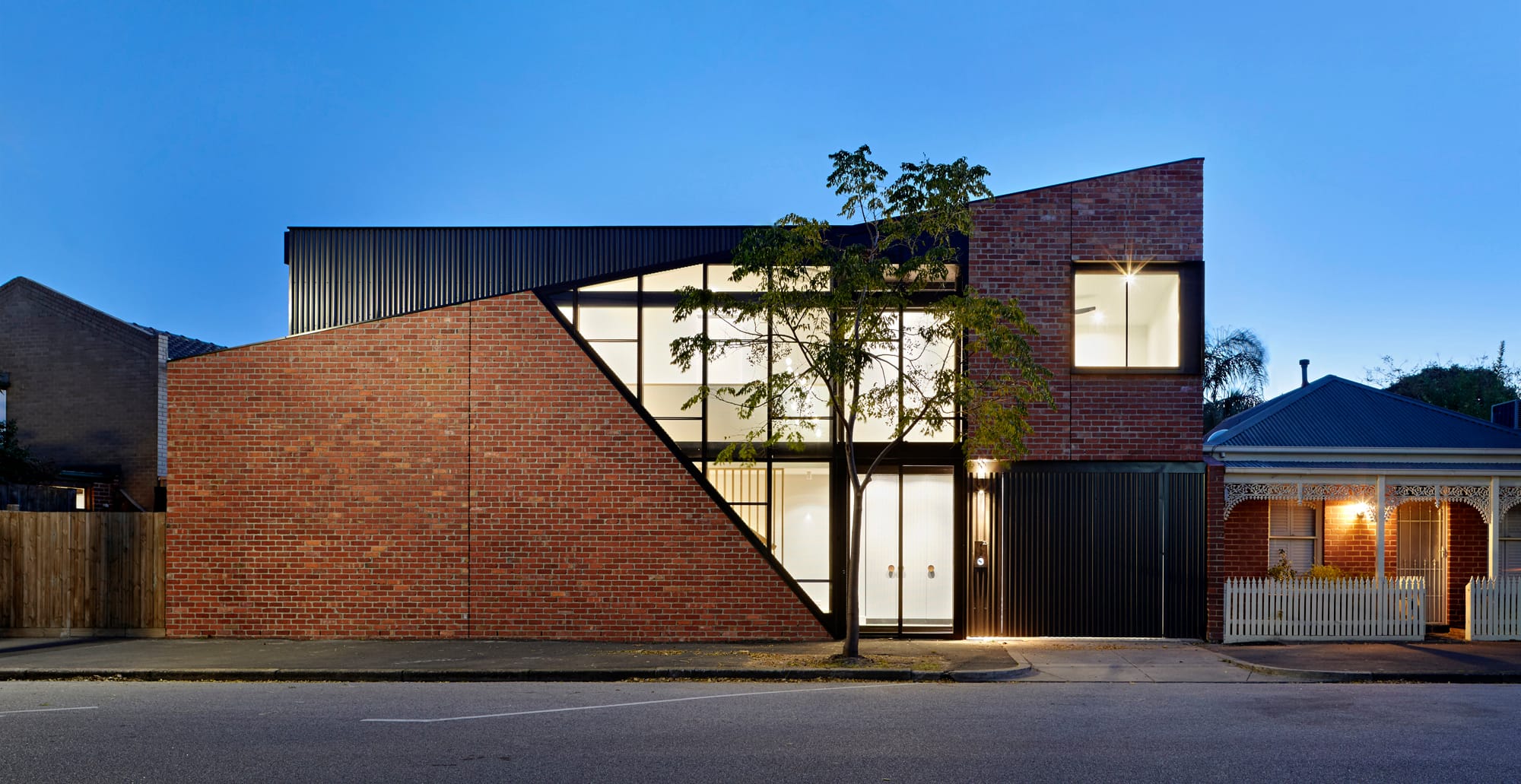 Contemporary brick house with angular roofline, large glazed facade, and warm interior lighting at dusk.