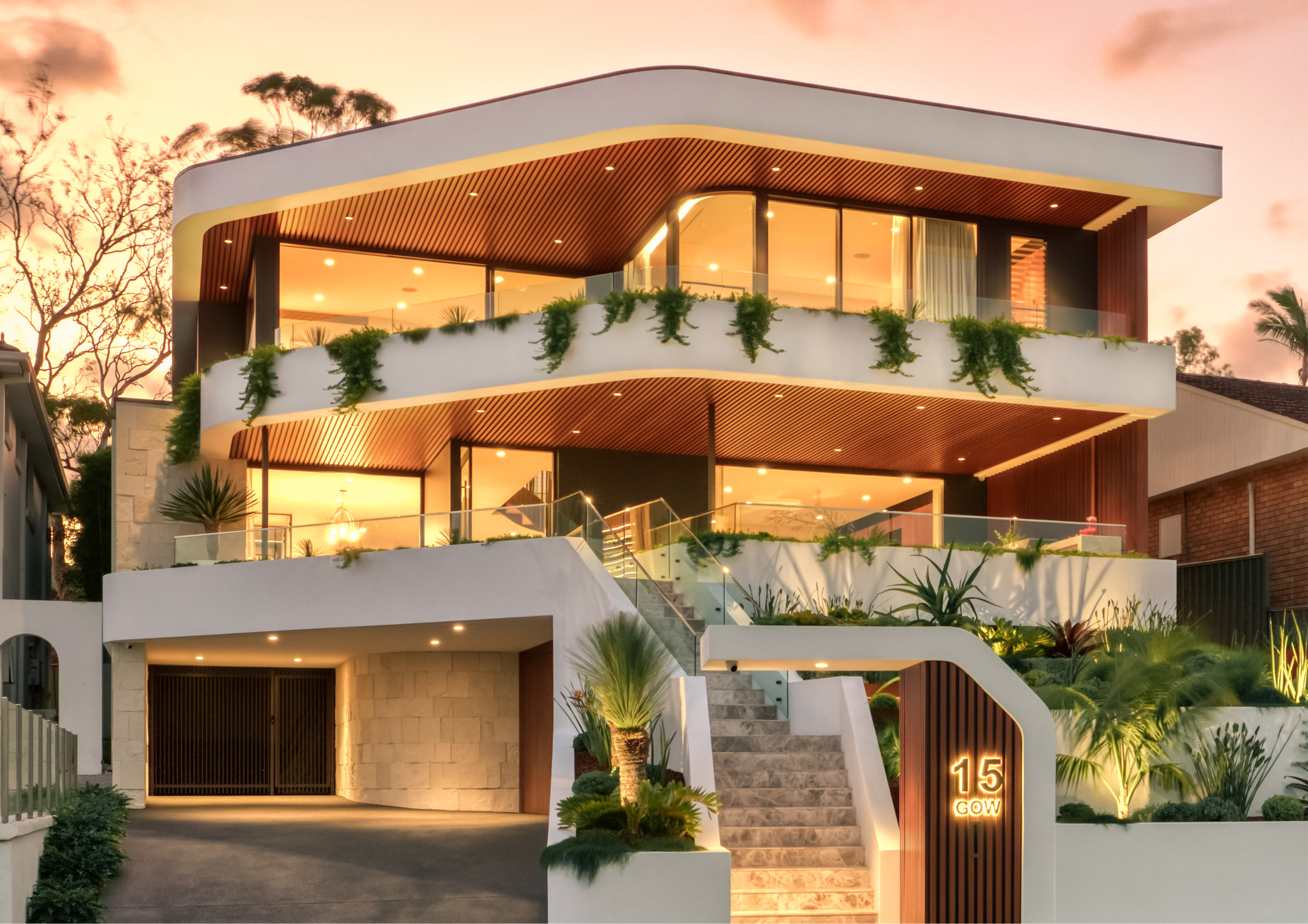 Contemporary multi-storey residence with curved white concrete forms, expansive floor-to-ceiling glazing, timber-lined soffits, and lush landscaped terraces illuminated at sunset.